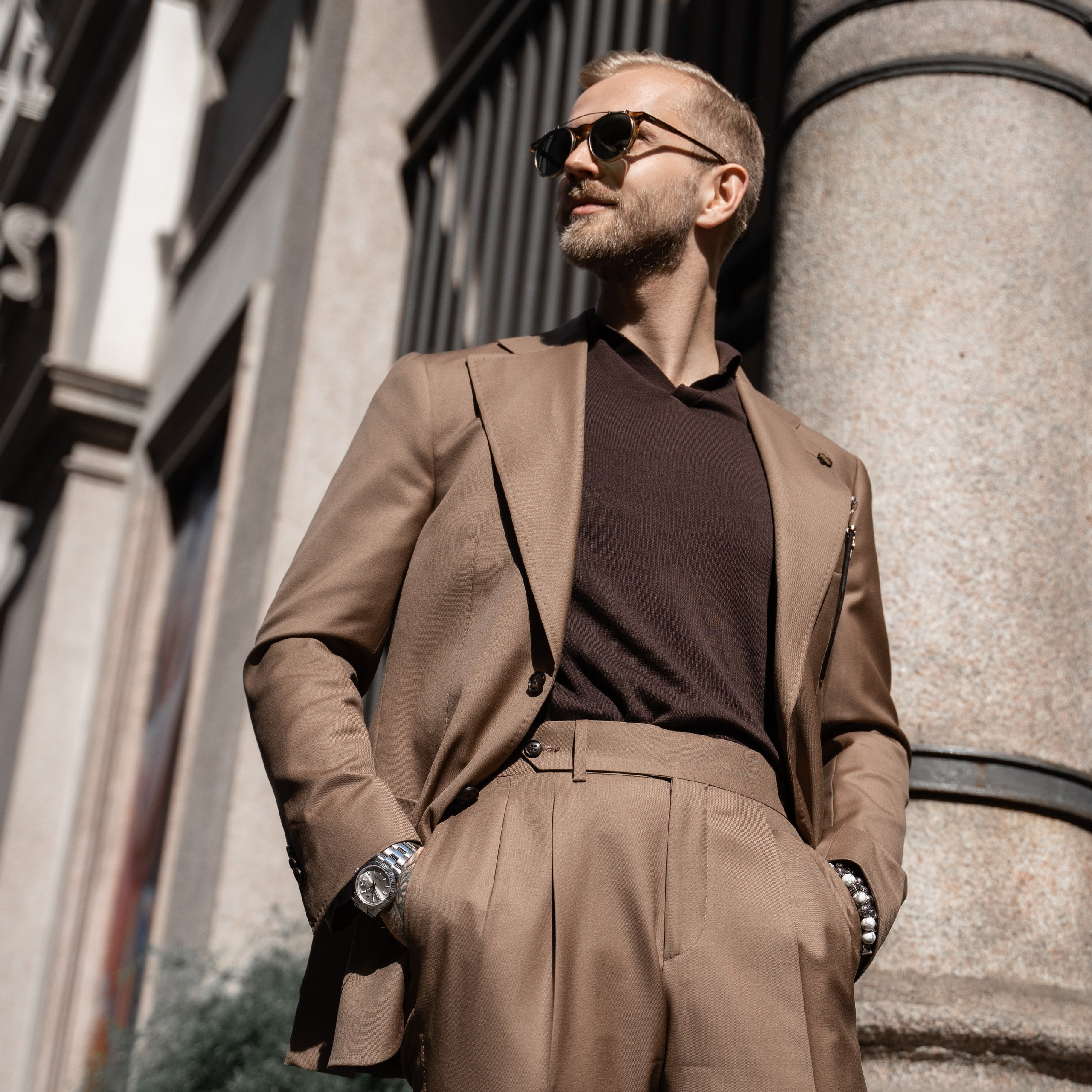 Editorial fashion shot of a confident man in a tan suit and sunglasses, leaning against a stone column in Milan. Men’s fashion photography, street portrait
