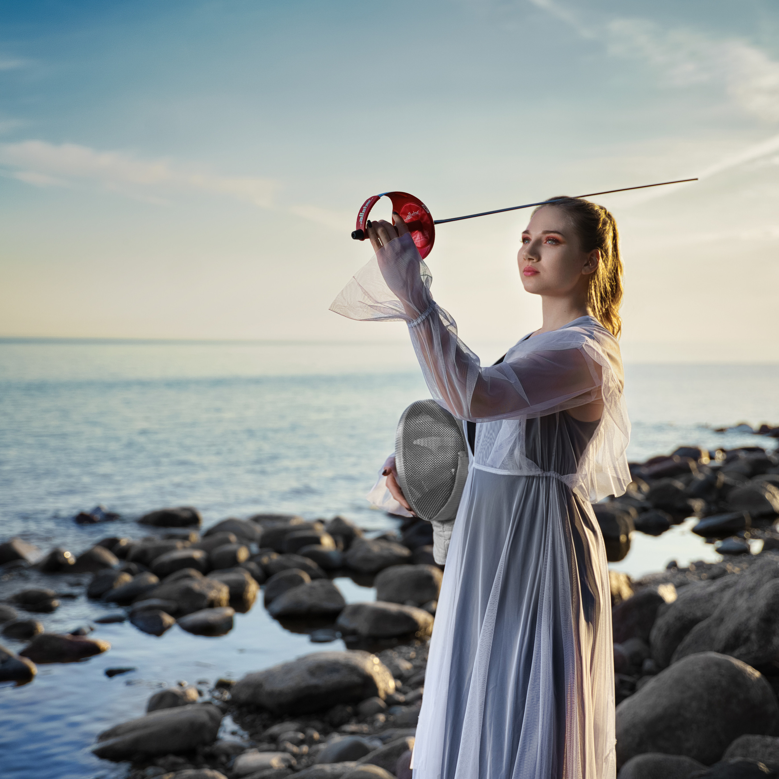Woman in a white dress holding a fencing foil by the sea