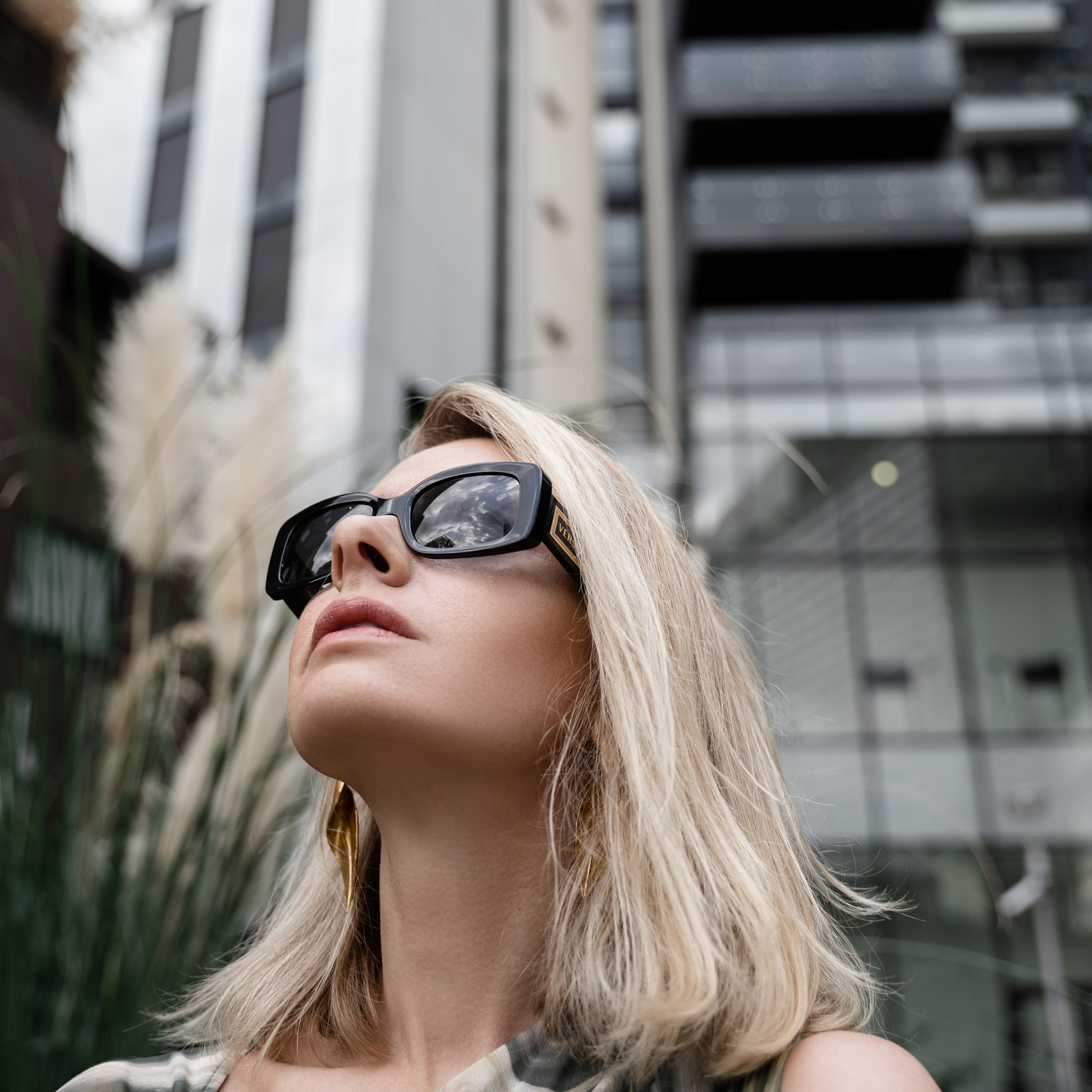 "Woman in stylish sunglasses looking upwards with modern buildings and pampas grass in the background