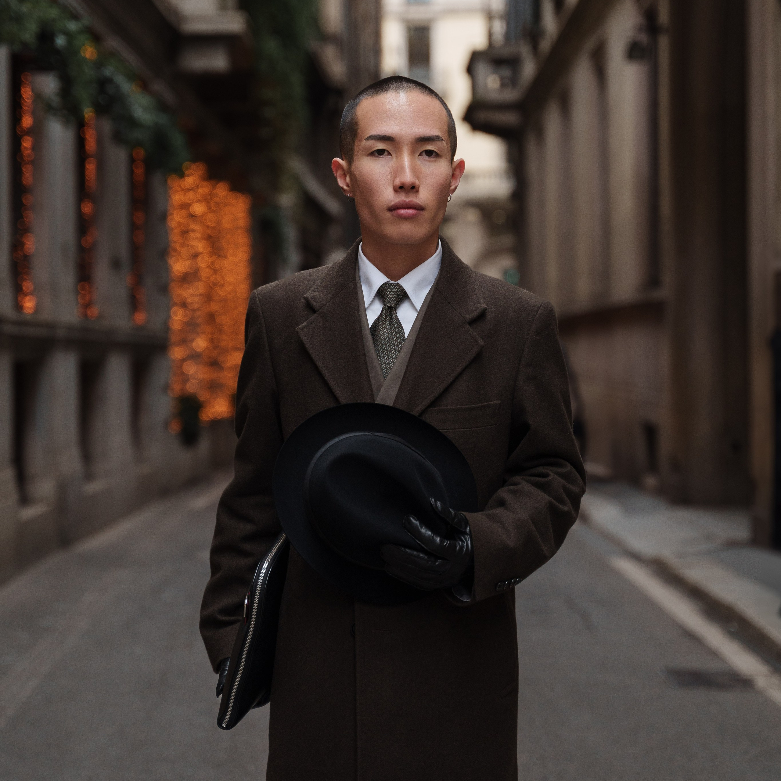 Stylish Asian man in a brown coat holding a black hat, standing on a narrow Milanese street with festive lights in the background