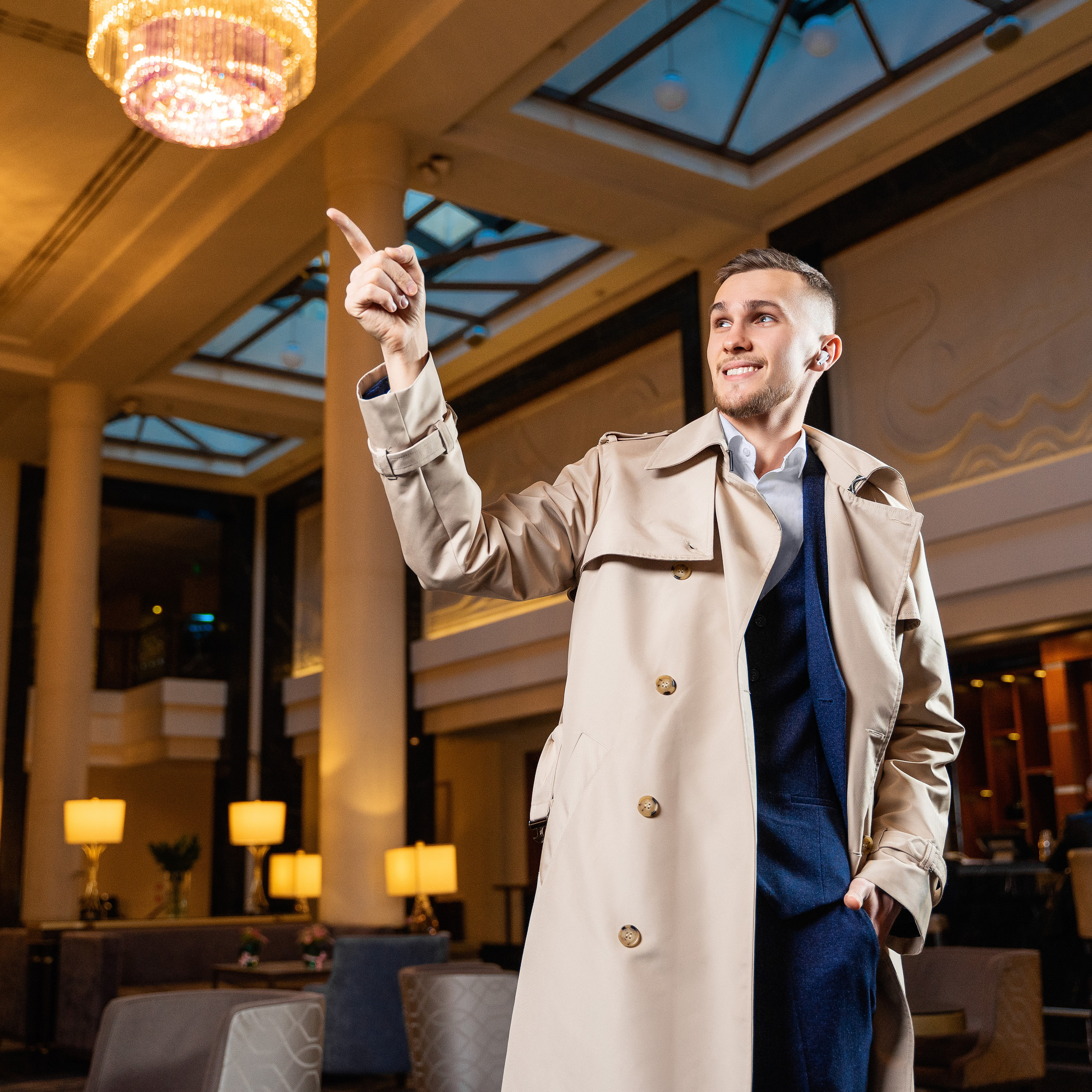 Charming man in trench coat raising finger to hail a taxi in opulent hotel entrance