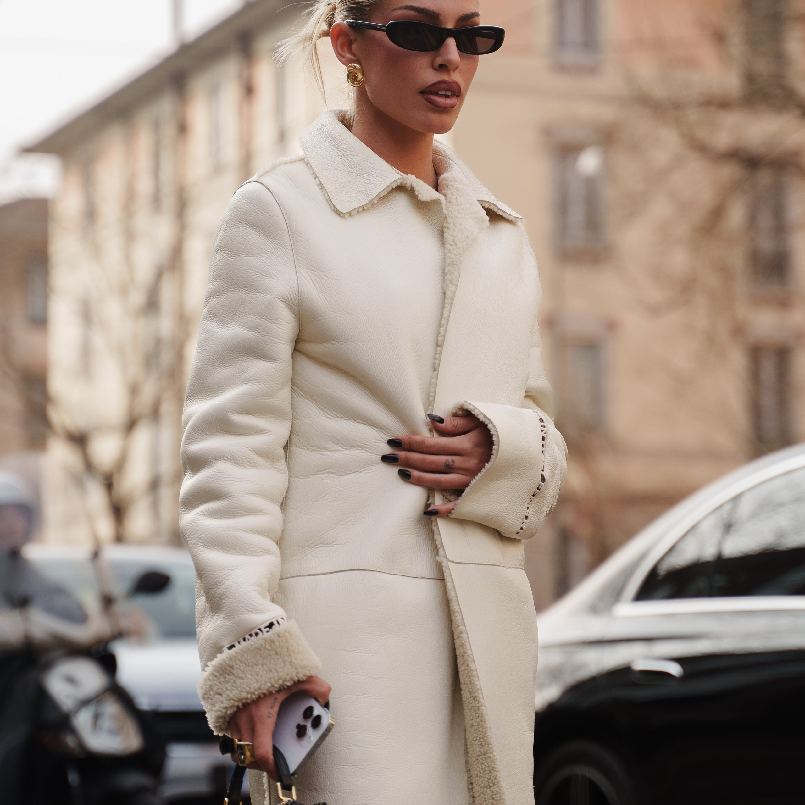Blonde woman in a white shearling coat holding a small black handbag, gazing to the side