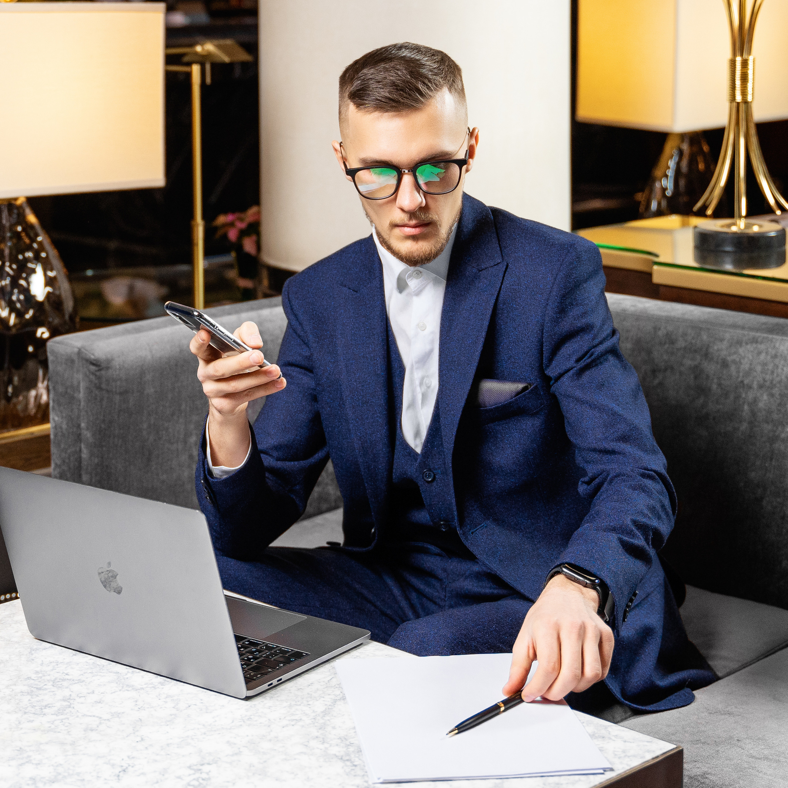 A focused businessman in a sharp blue suit is engaged in multitasking, with one hand holding a smartphone and the other holding a pen over a notepad, sitting at a polished table with a MacBook and sophisticated decor, indicating a luxurious workplace setting