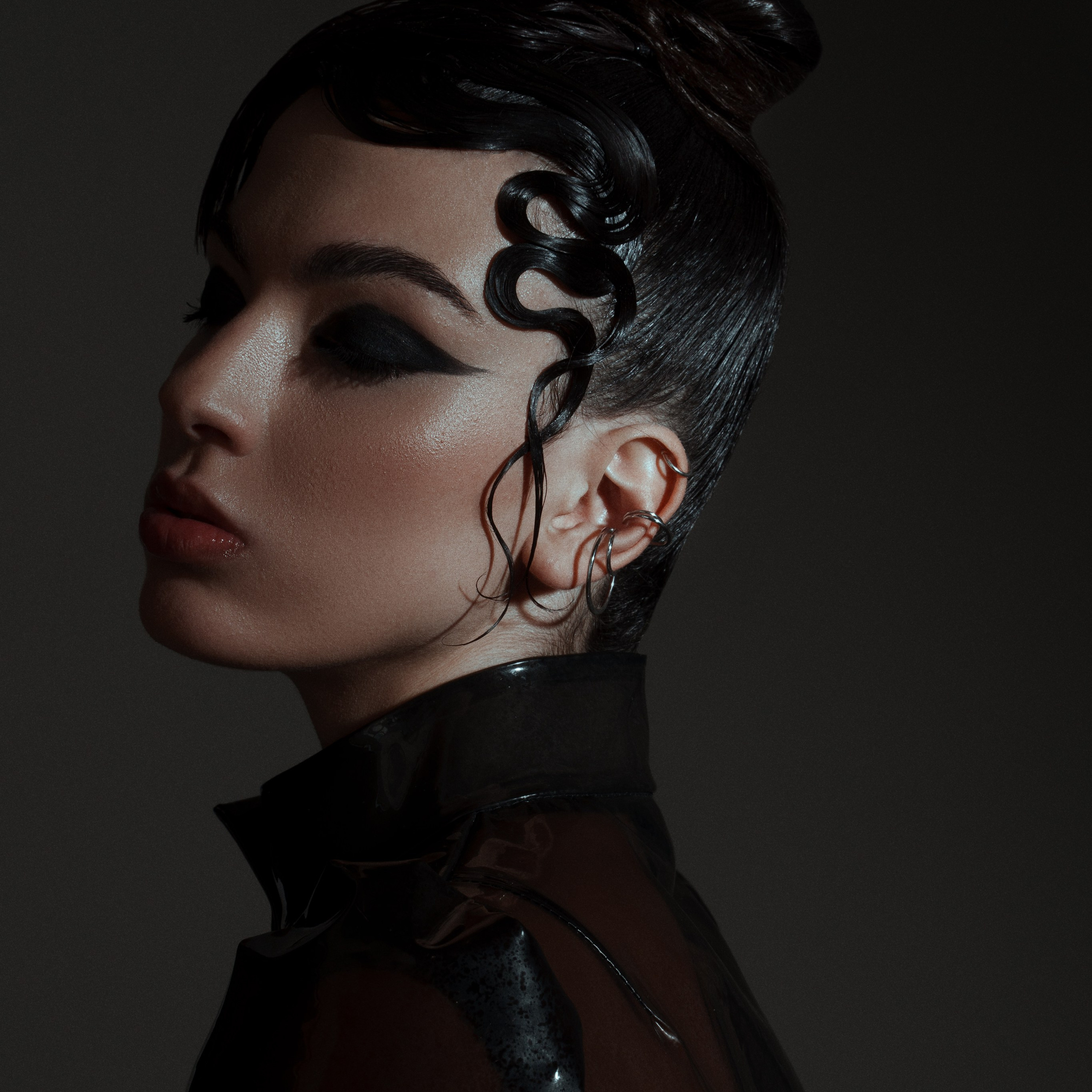 Avant-garde beauty portrait of a woman with bold black eyeliner, sculpted wet-look hair, and a high-fashion latex coat, set against a dark moody background