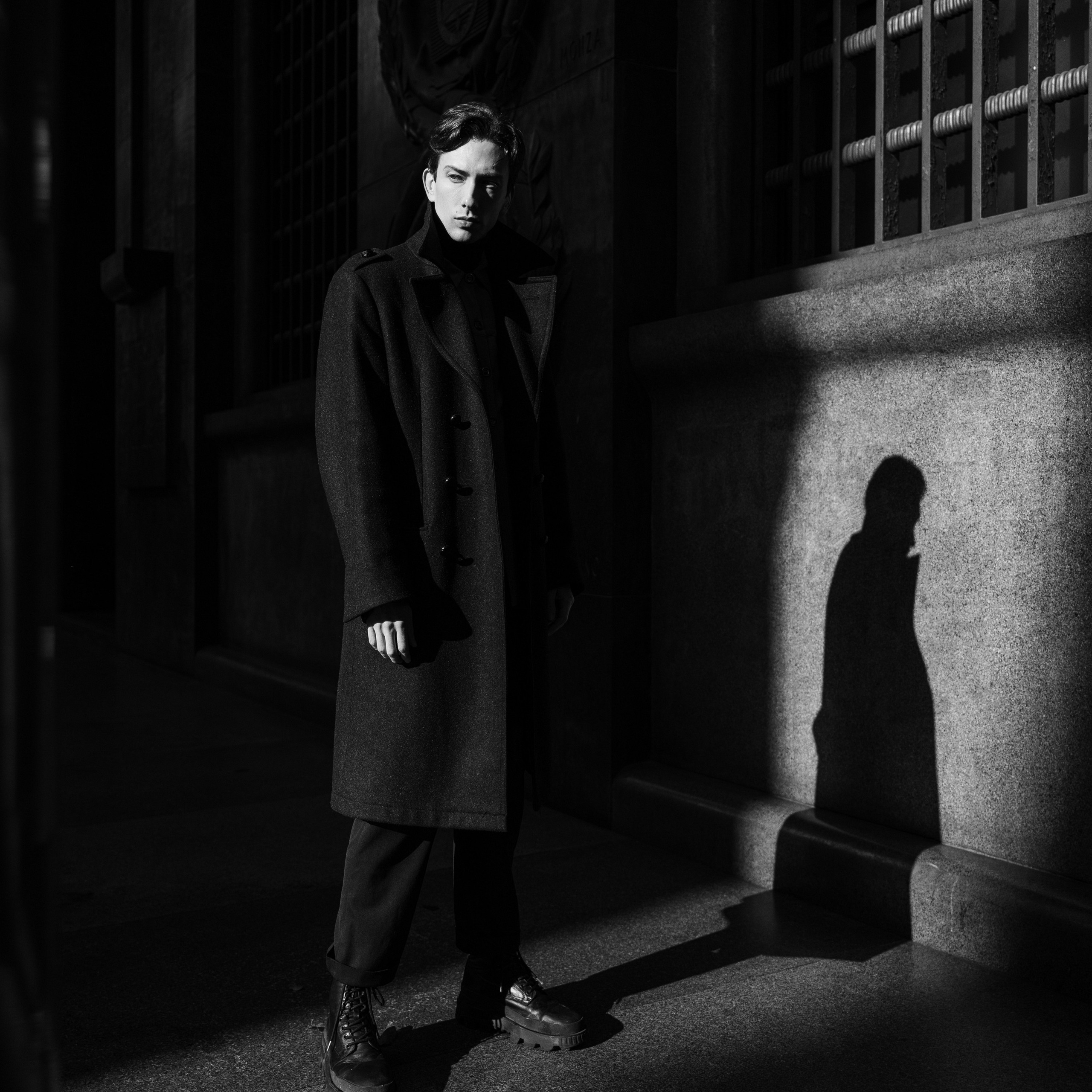 Monochromatic image of a stylish young man posed in a striking beam of sunlight, casting a long shadow, against the classic architecture of a city building, creating a dramatic and moody atmosphere