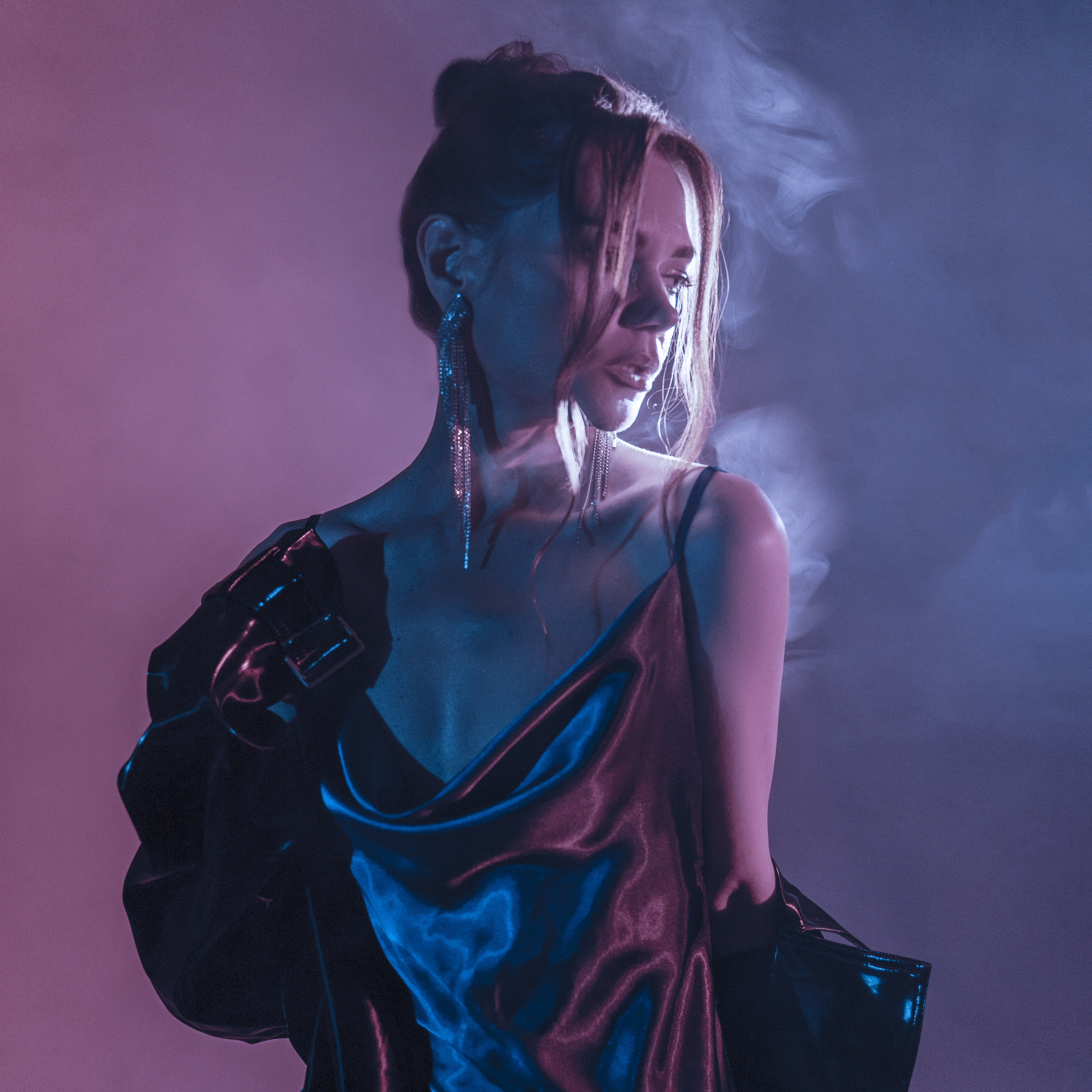 Elegant woman in a red silk dress with sparkling earrings, holding a jacket over her shoulder with a smoky backdrop