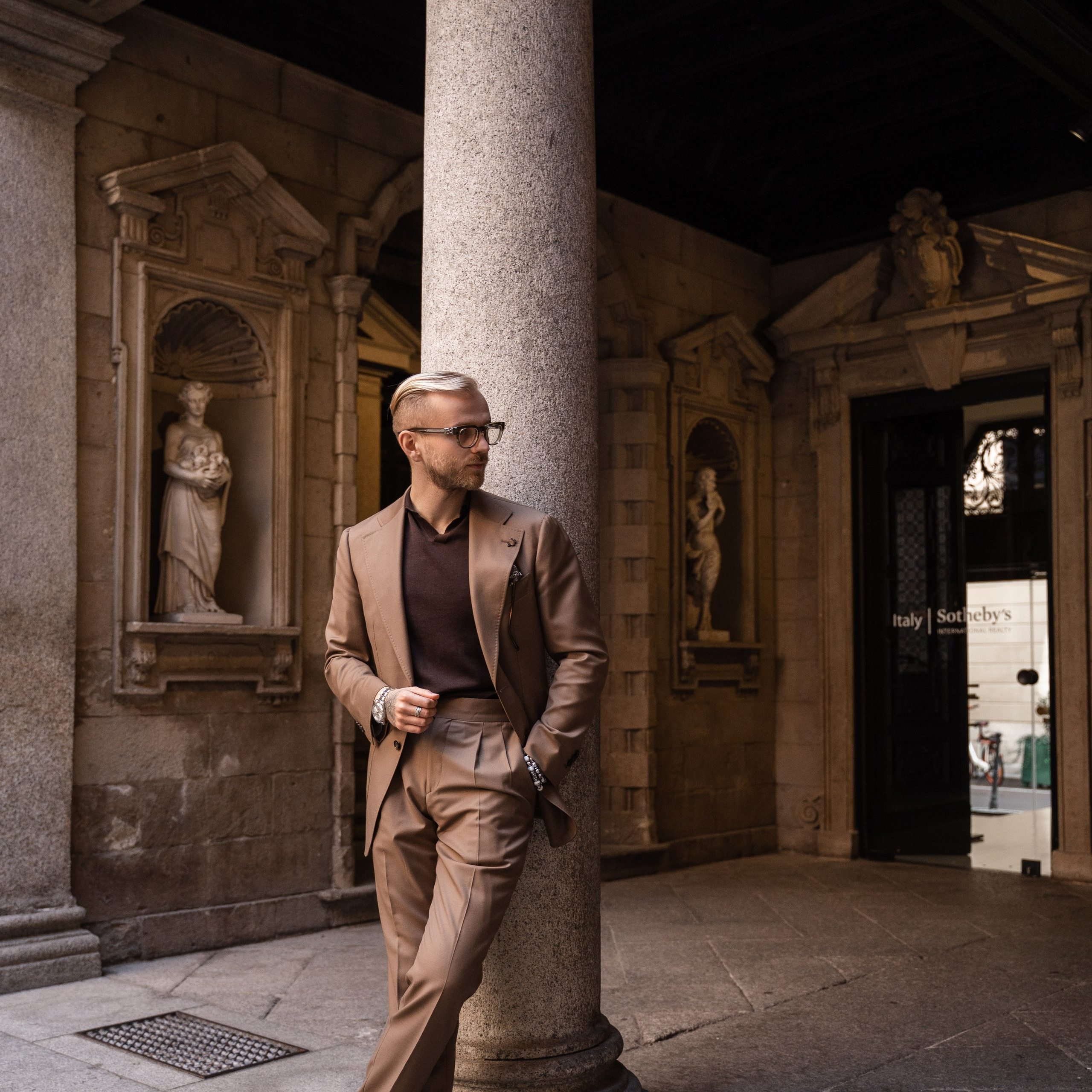 Elegant man in a beige suit leaning against a stone column in a historic Milanese courtyard
