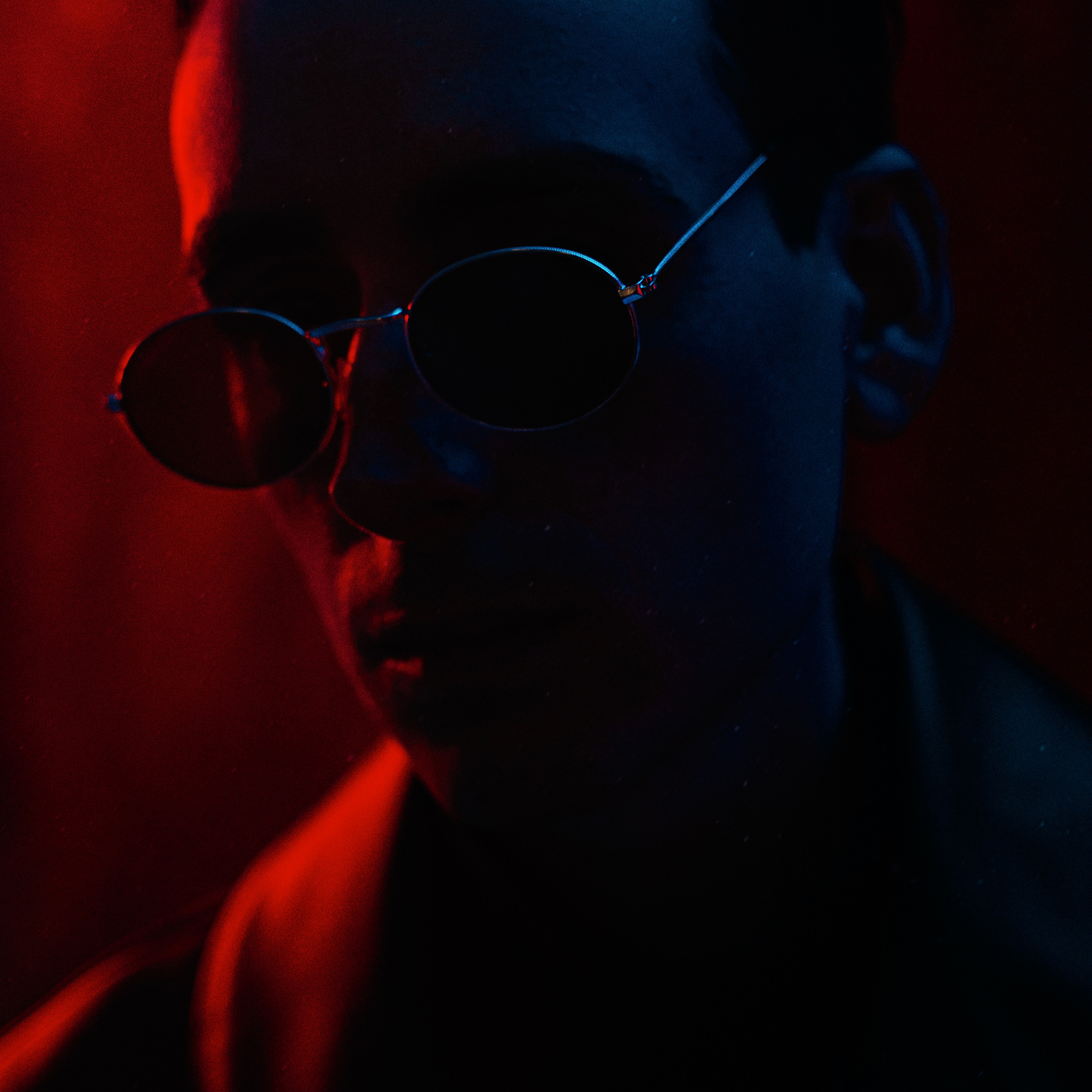 Dark moody portrait of a man wearing round sunglasses with red lighting highlighting his face