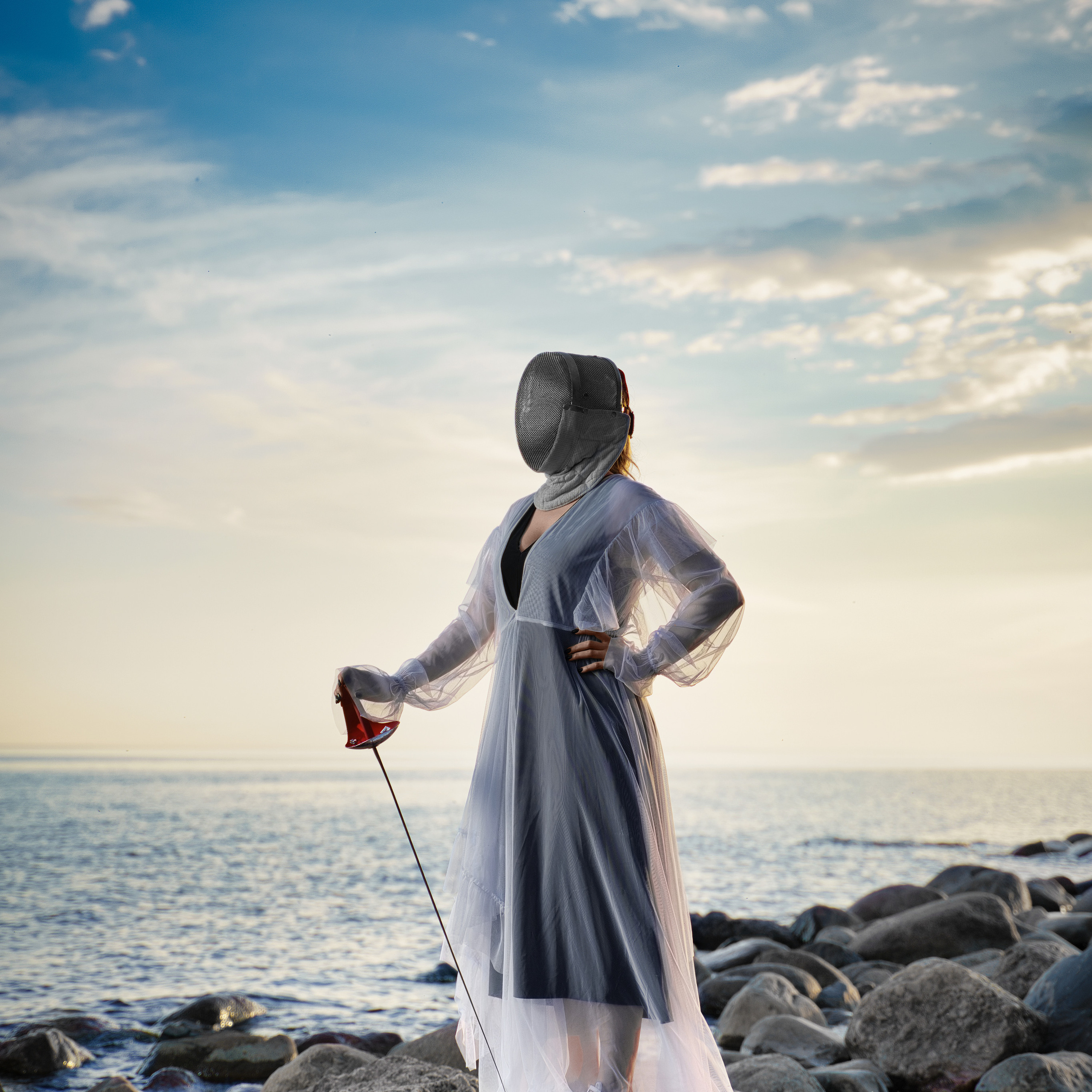 Figure in white translucent gown and fencing mask standing on rocks at seaside during sunset