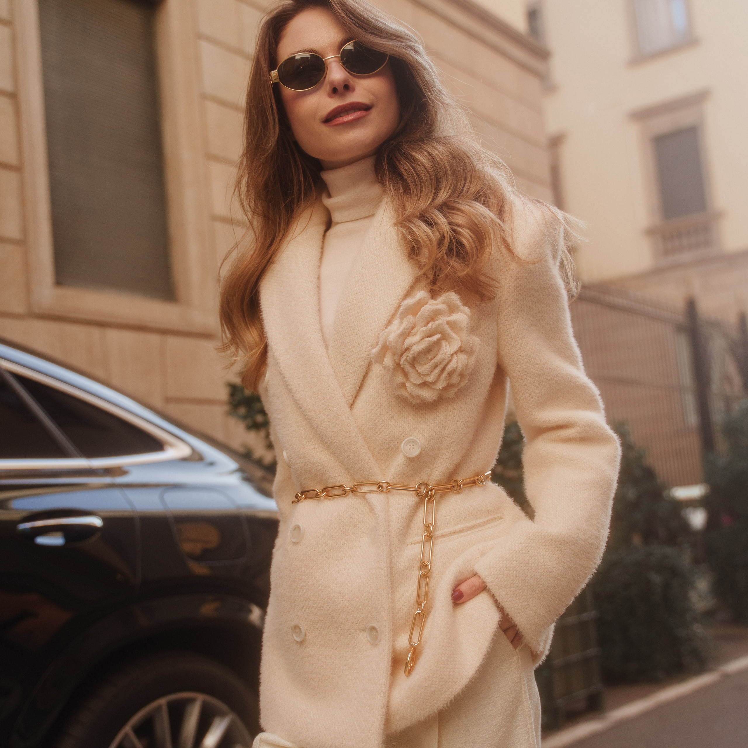 Elegant woman in a white wool coat with gold accessories, sunglasses, and a luxury car in the background on a Milan street