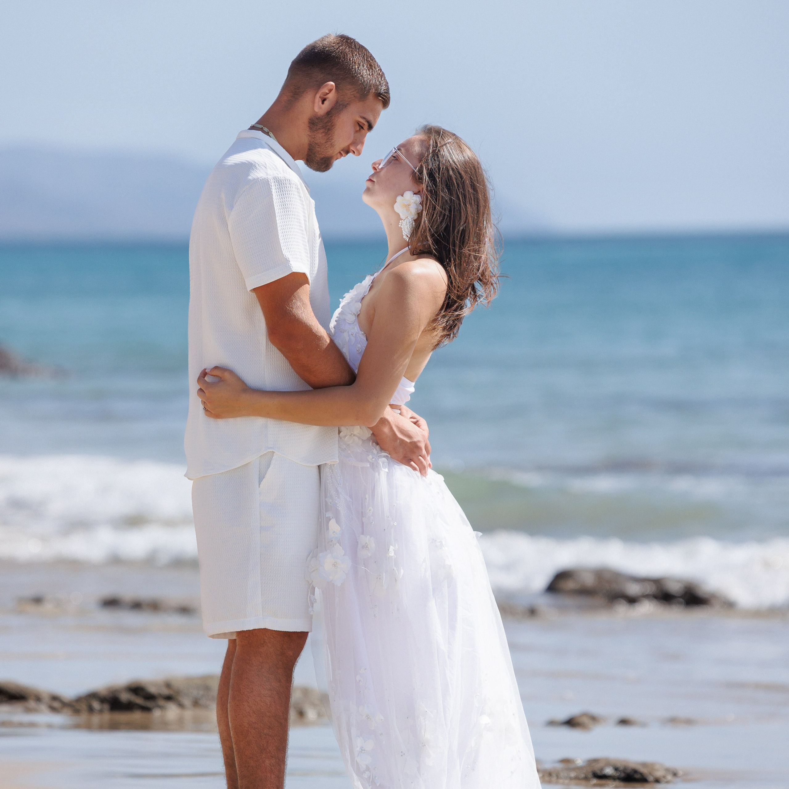 Couple holding hands while walking barefoot along the sandy beach at dawn, with soft ocean waves rolling in.