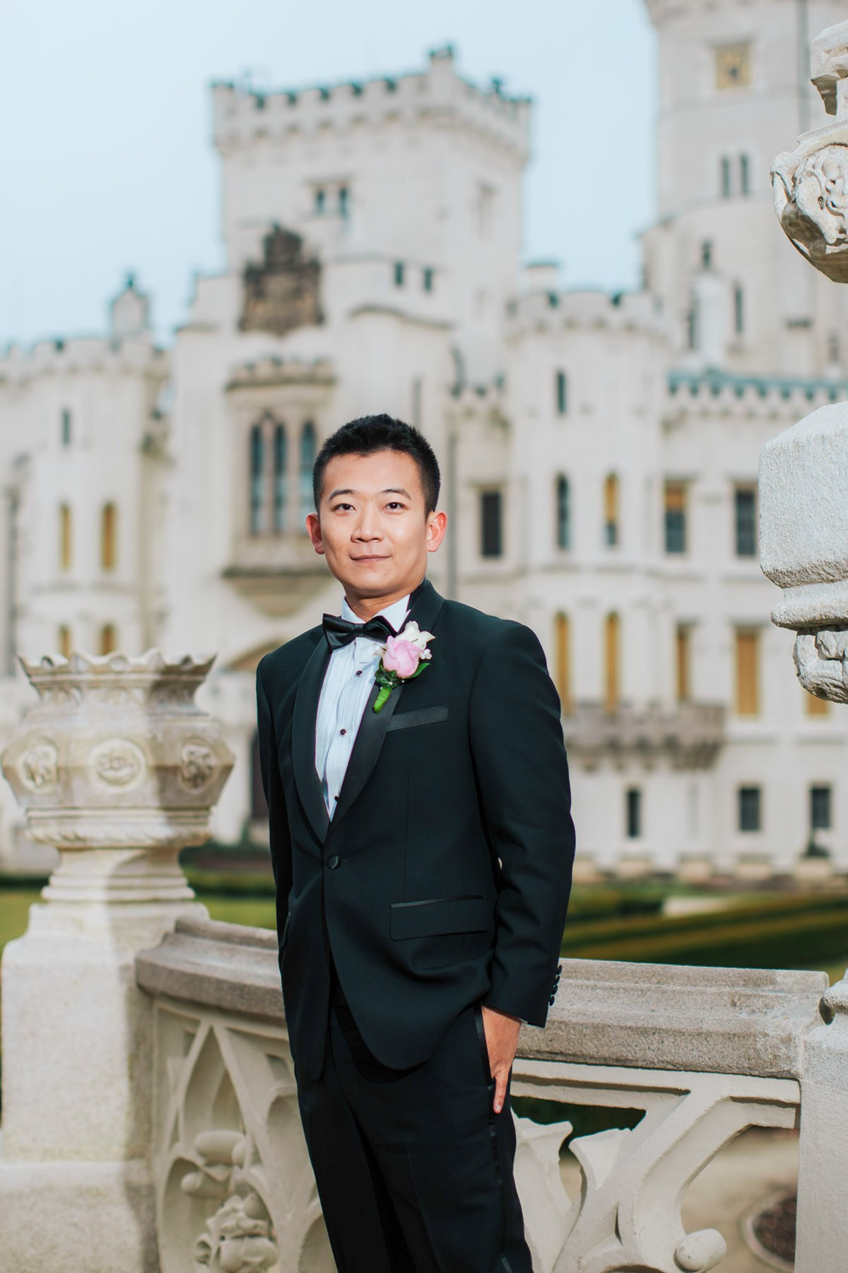 A smling, tuxedo wearing groom poses for a portrait against the backdrop of the impressive Castle Hluboka during his destination wedding.