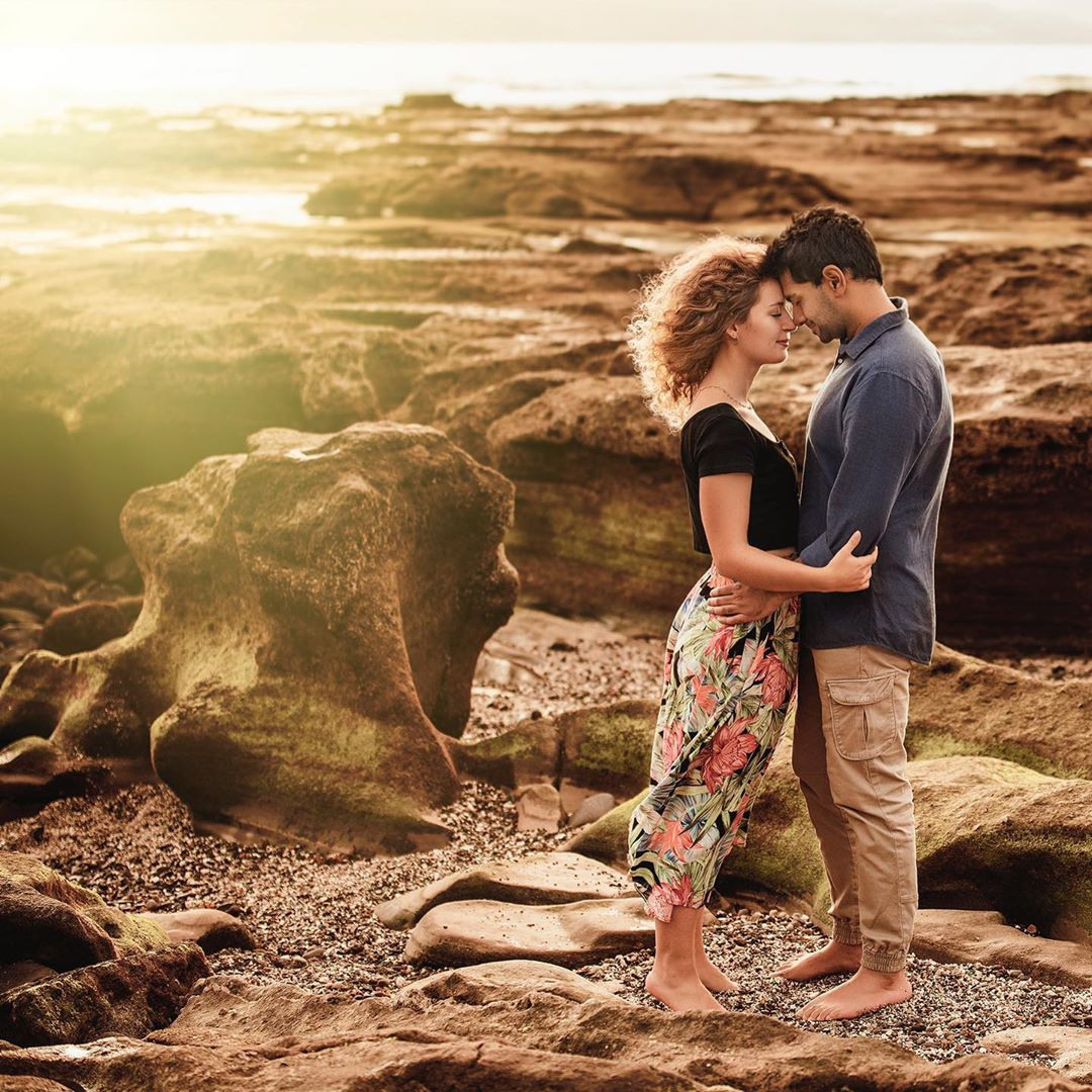 An engaged couple standing on rocks at sunset.