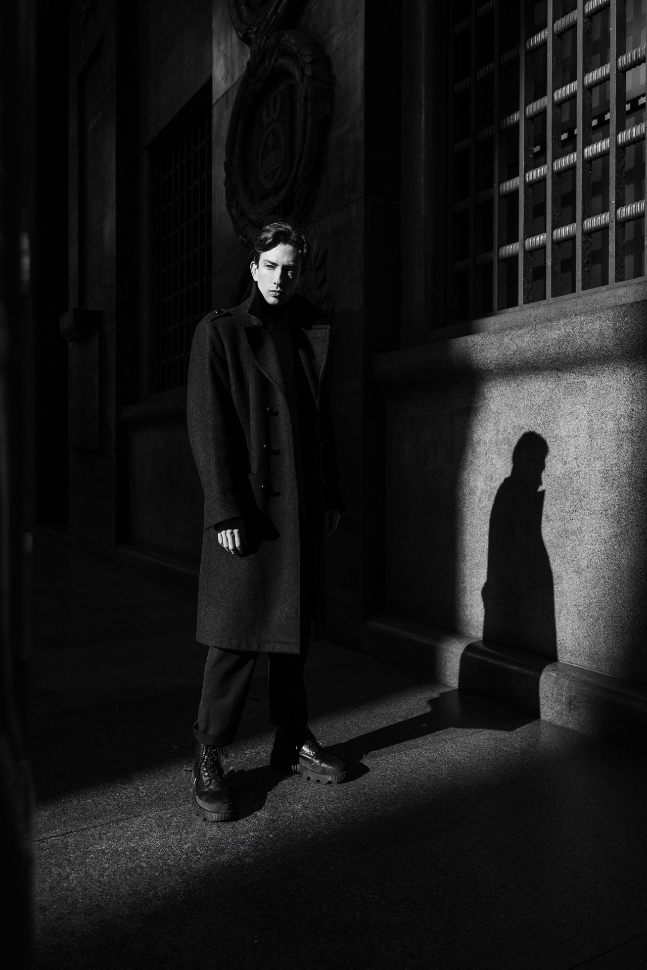 Male model in long dark coat posing by textured stone wall in Milan
