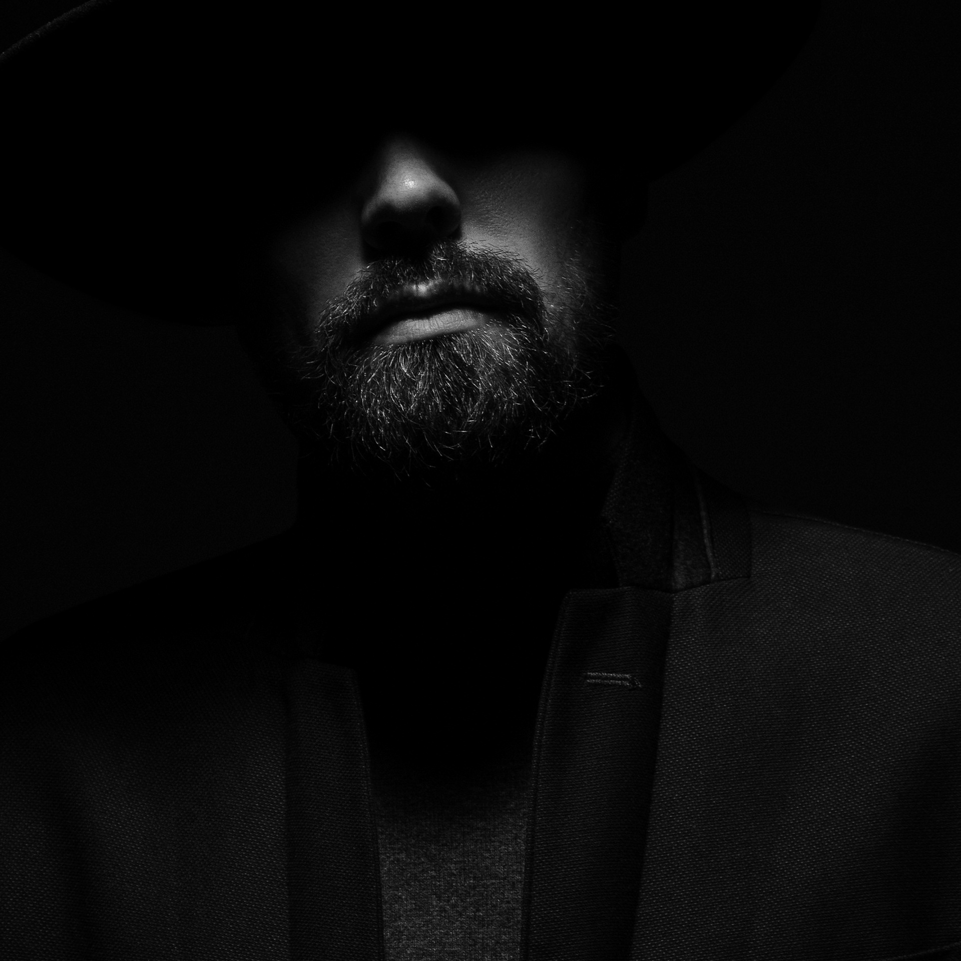 Close-up of a man's face with a beard, wearing a hat, shrouded in shadows
