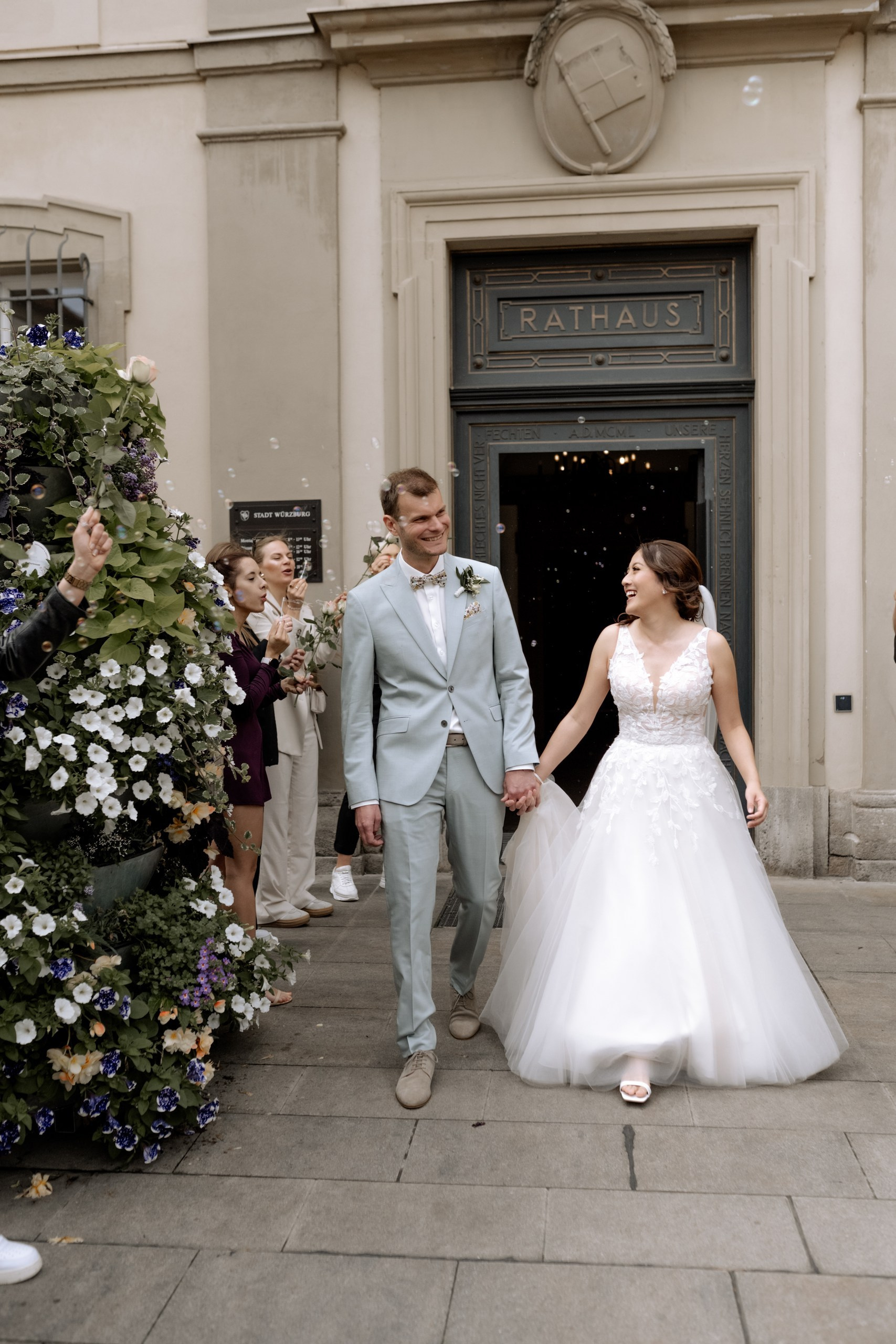 Das frisch vermählte Paar verlässt Hand in Hand das Standesamt Würzburg, umjubelt von Freunden und Familie. Luftschlangen und Konfetti sorgen für festliche Stimmung.