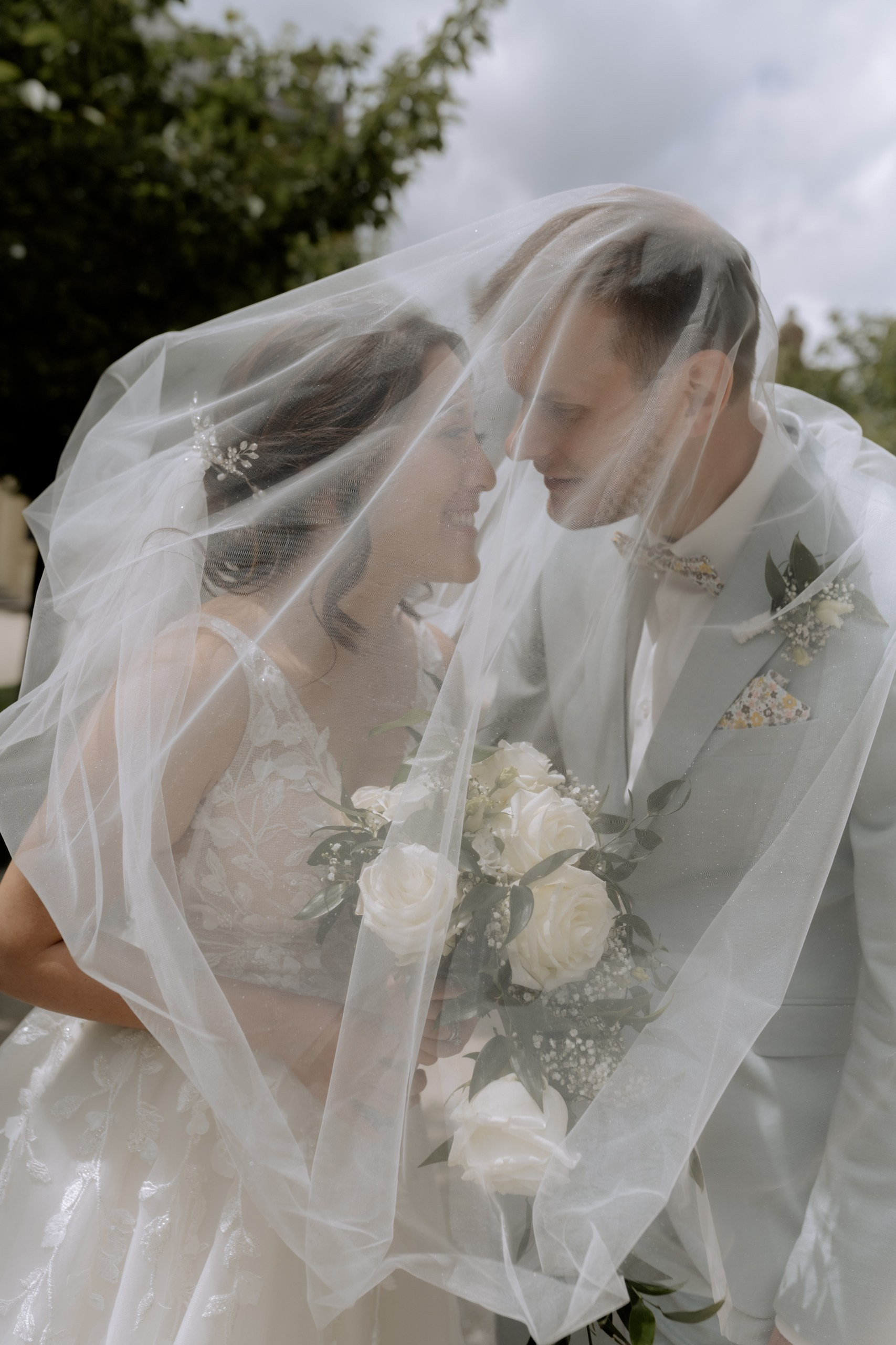 Die Braut in einem eleganten weißen Kleid steht vor einer alten Steinmauer, der Schleier weht im Wind. Ihr Blick ist verträumt und glücklich.
