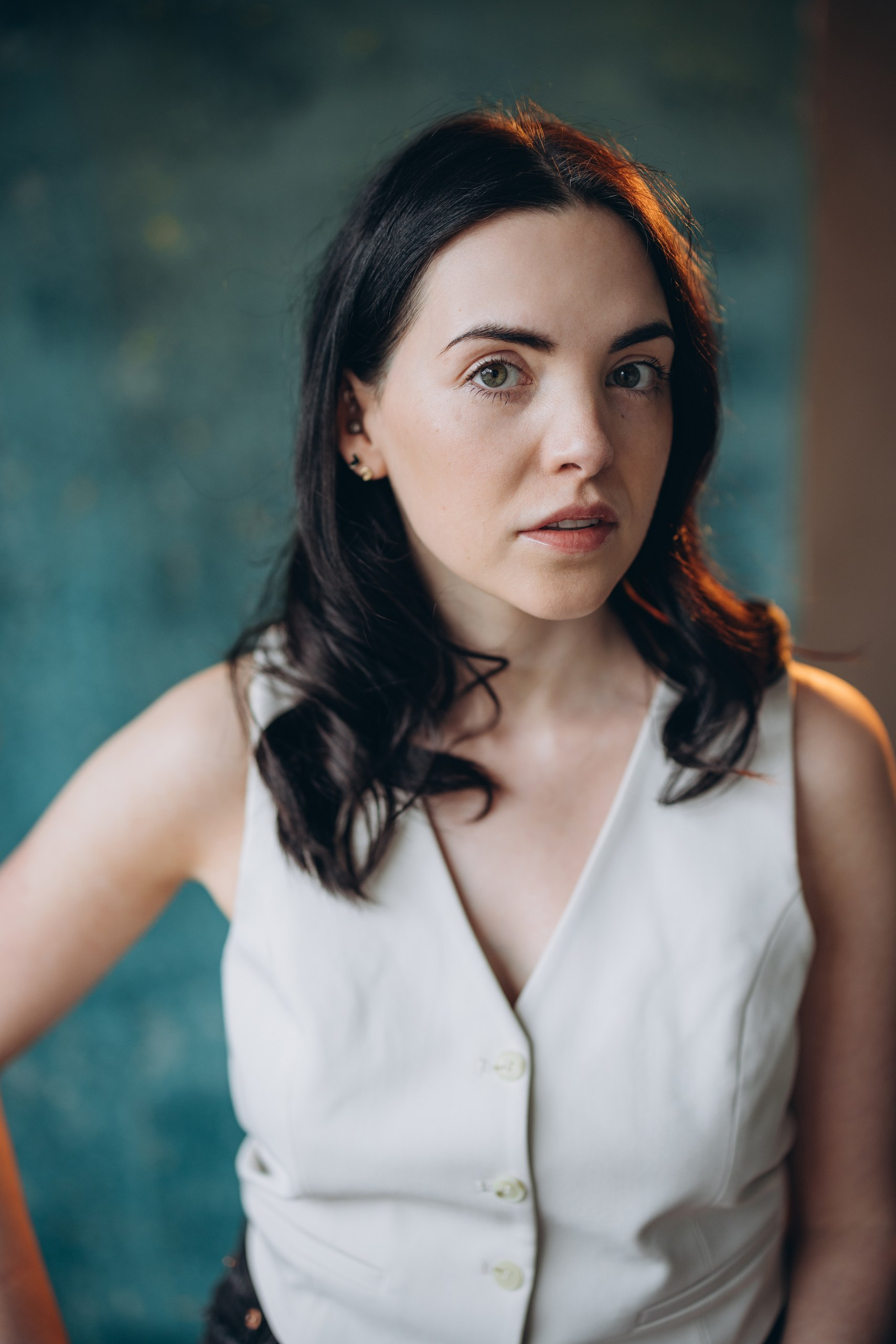 Actor headshot of a serious woman with dark hair wearing a sleeveless white blouse, sitting against a dark background