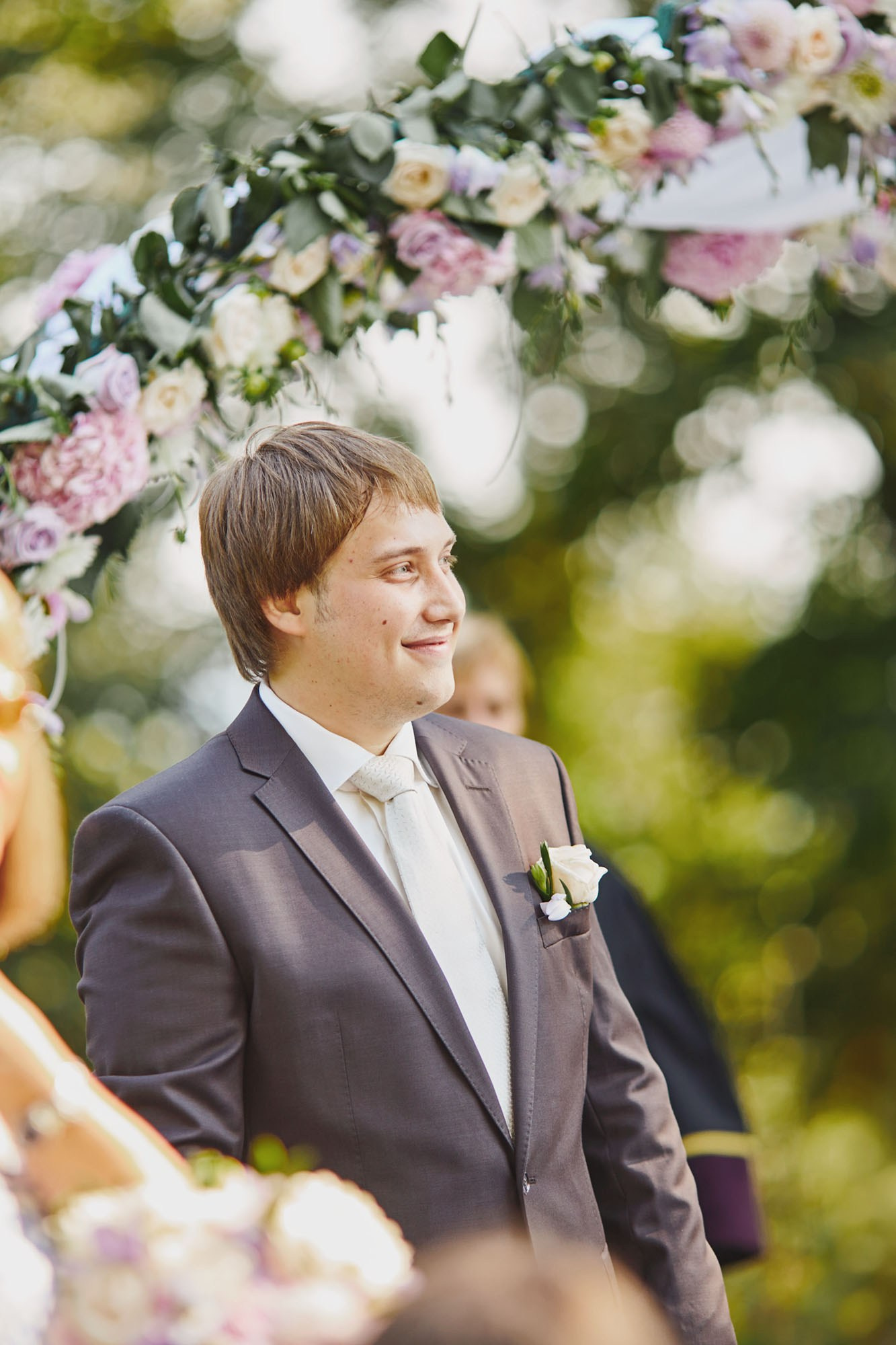 A smiling groom, wearing a dark brown suit, smiles at the first site of his bride as he stands under a floral arch during his wedding ceremony.