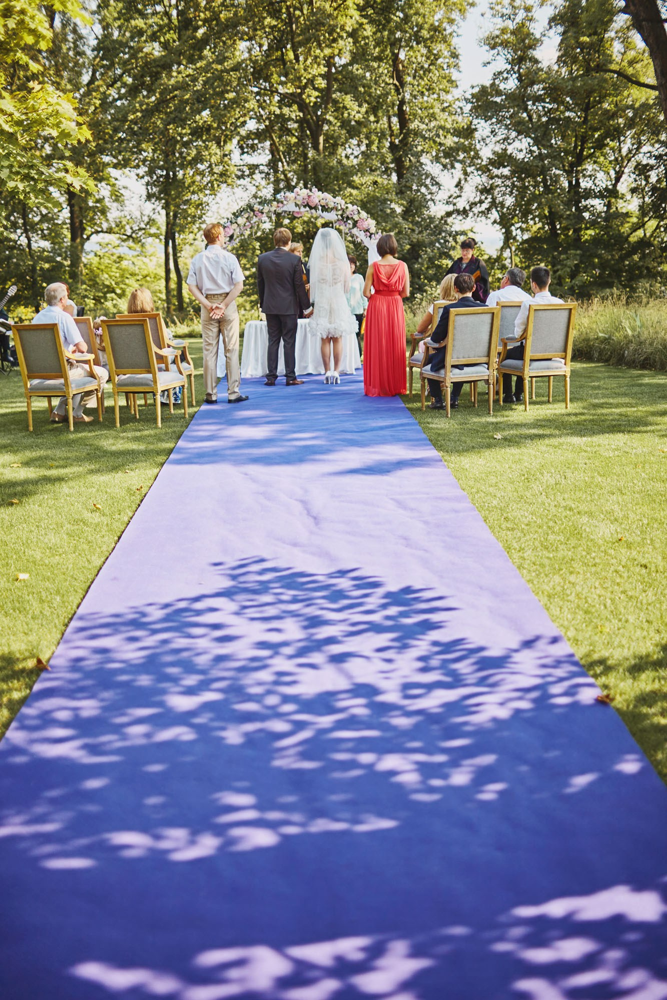 Wedding party on French blue carpet under floral arch at chateau for vow exchange.