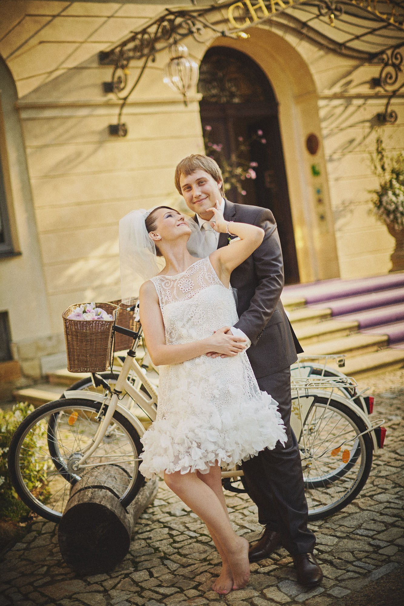 Young newlyweds share a tender moment, caressing each other beside vintage bikes at the grand entrance of Chateau Mcely.