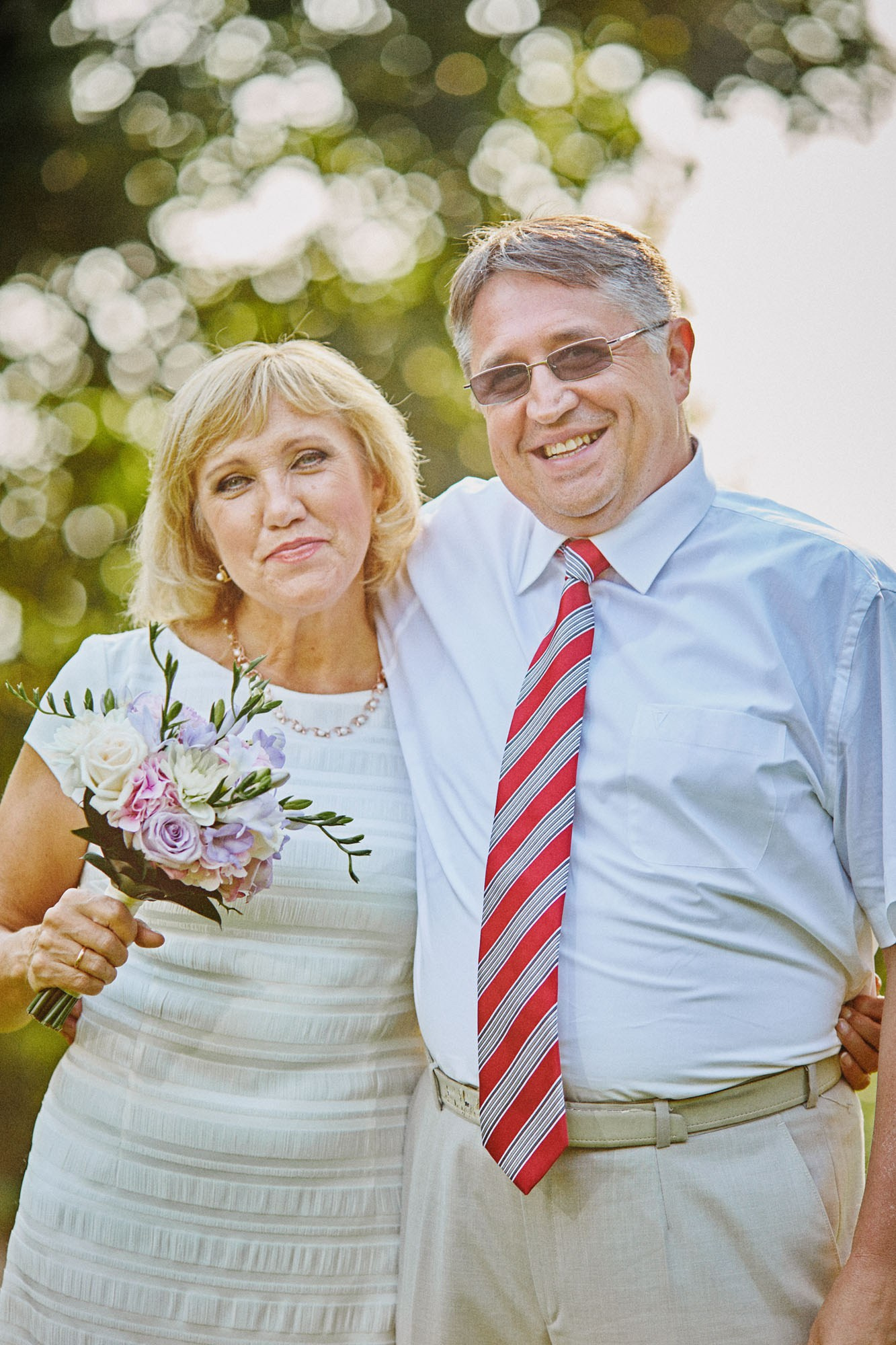 The groom's parents cuddle up for a photo as the mother holds a bouquet of flowers following their son's beautiful summer wedding.