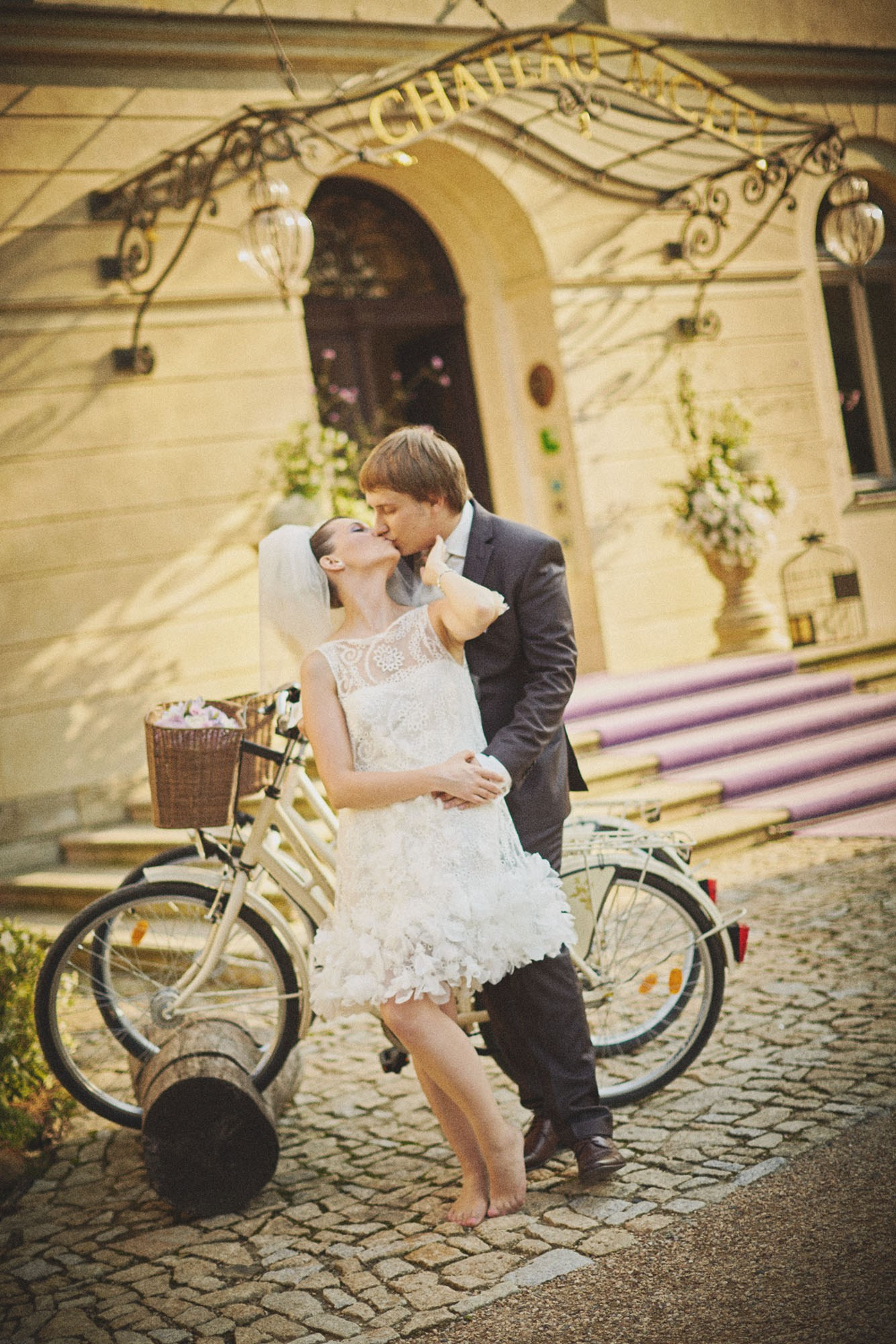 Young newlyweds kiss beside vintage bikes at the grand entrance of Chateau Mcely.