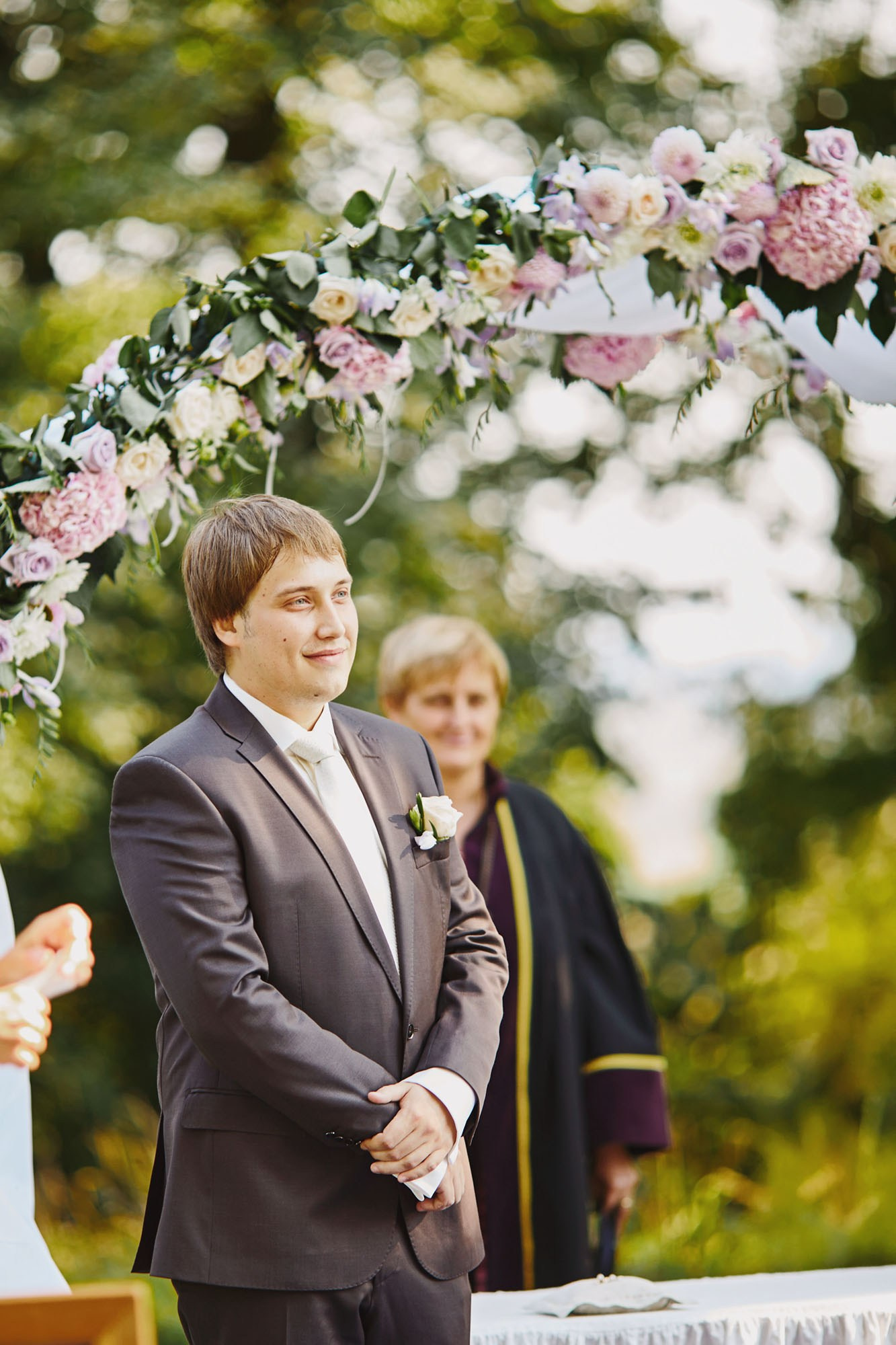 Groom in dark brown suit under floral arch awaiting bride at outdoor wedding.