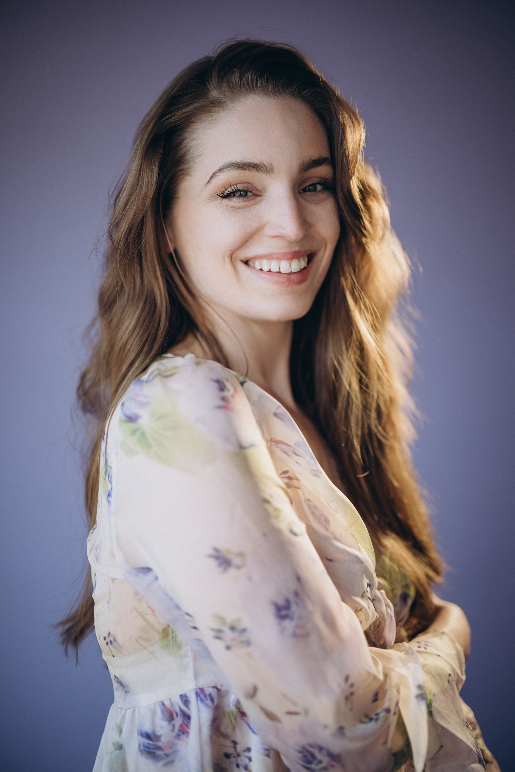 Actor headshot of a smiling woman with long hair, wearing a light floral blouse against a purple background