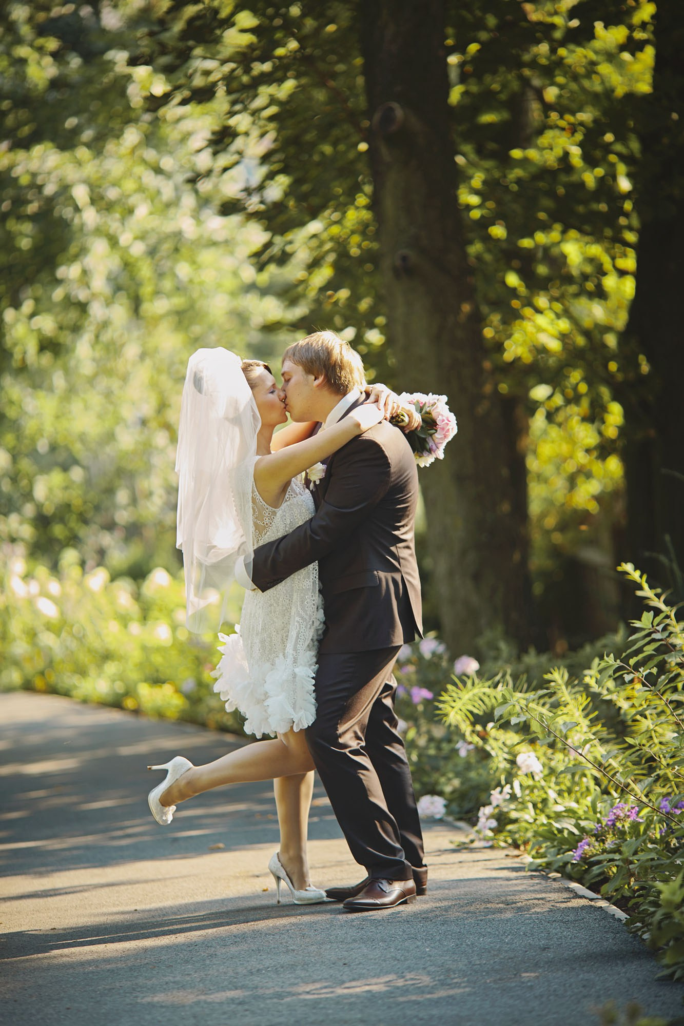 Newlyweds kiss under the trees on a flower-lined path at Chateau Mcely, capturing a picturesque moment after their outdoor wedding ceremony.