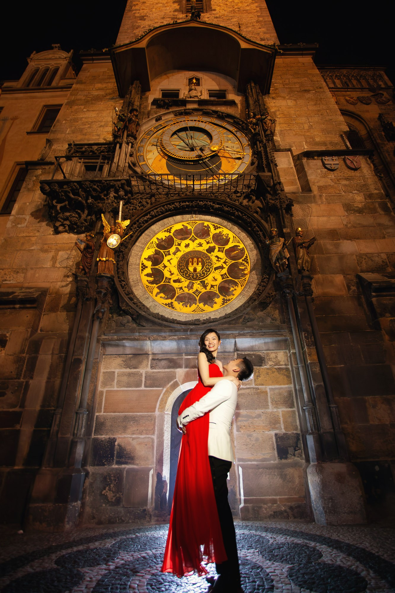 A smiling Hong Kong woman wearing a stylish red evening dress and is picked up and spun around underneath the historic Astronomical Clock in Prague during their night time love story photo session.