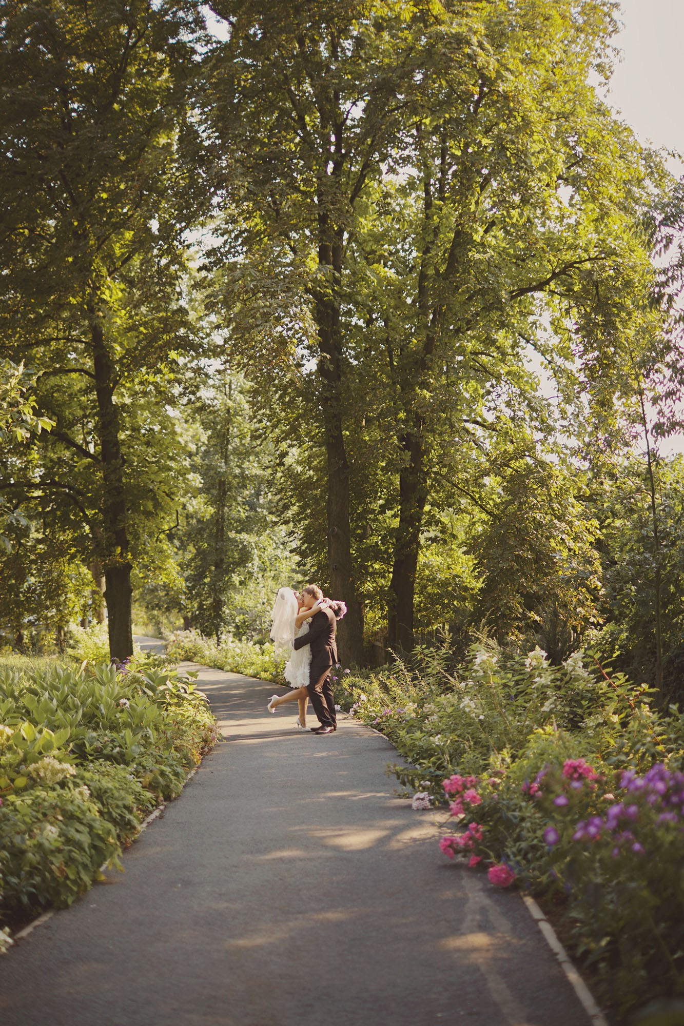 Newlyweds embrace under the trees on a flower-lined path at Chateau Mcely, capturing a picturesque moment after their outdoor wedding ceremony.