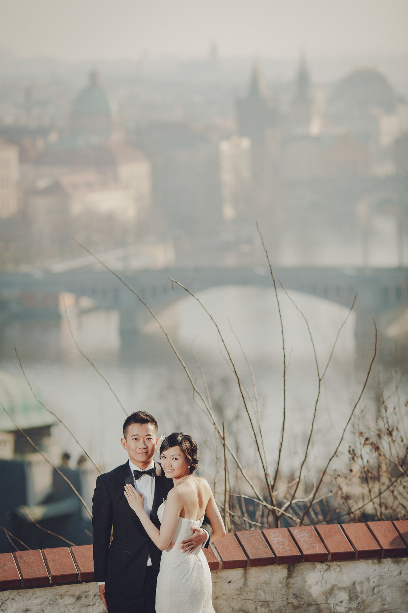A smiling Hong Kong couple, dressed in their wedding attire pose for a portrait high above the historic center of Prague.