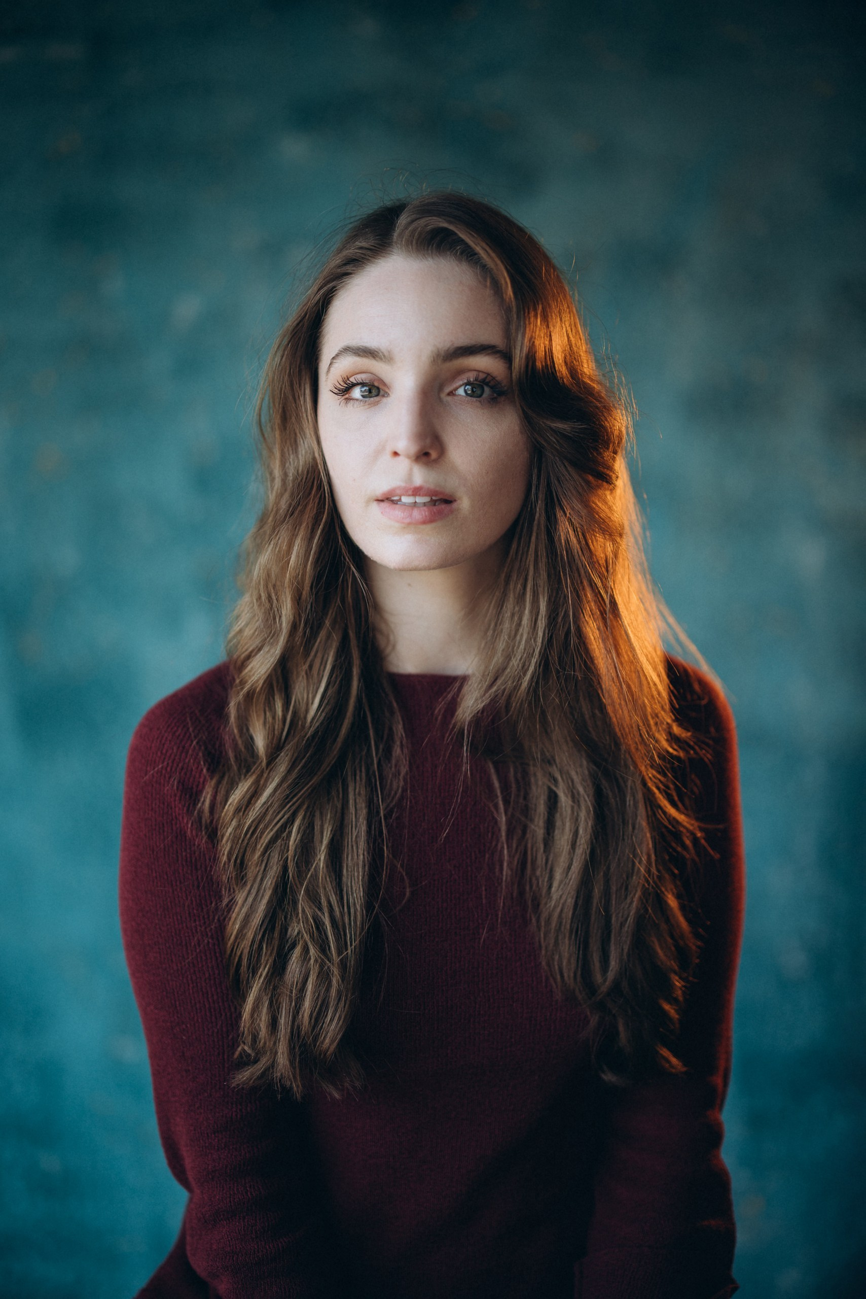 Actor headshot of a woman with wavy hair wearing a burgundy top, looking at the camera against a textured blue background