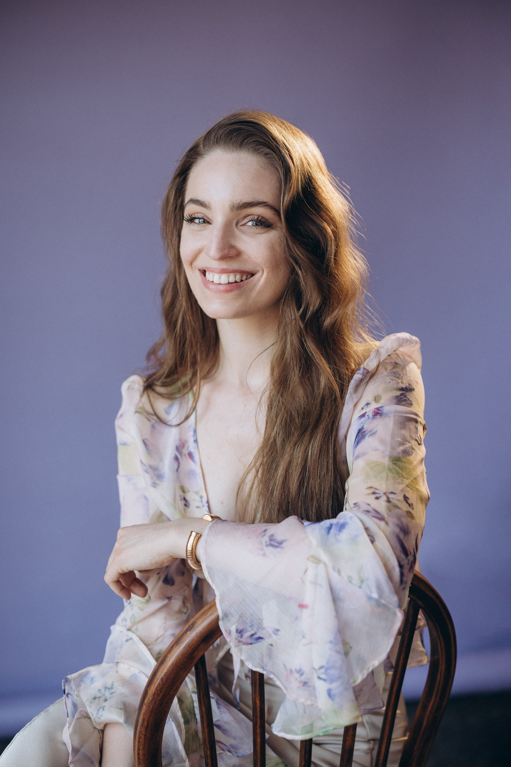 actor headshot of a smiling woman with long hair in a light floral blouse, sitting on a chair against a purple background