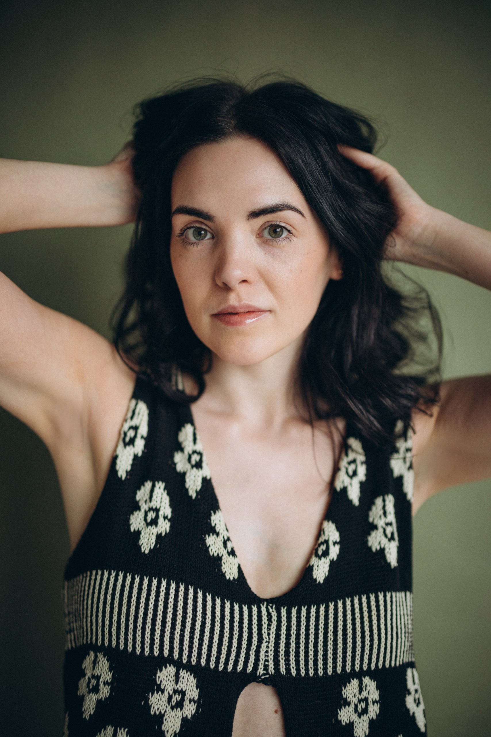 Actor headshot of a woman with dark hair wearing a patterned black dress, arms raised behind her head, looking into the distance