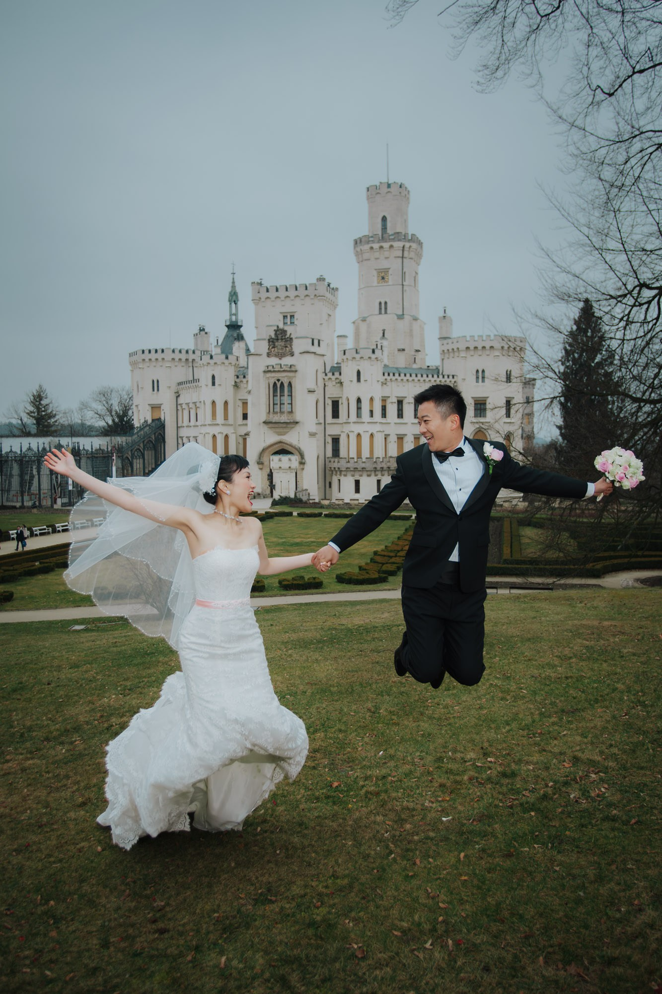 A smling Hong Kong bride & groom jump into the air against the backdrop of the famous State Chateau of Hluboka during their destination wedding in the Czech Republic.