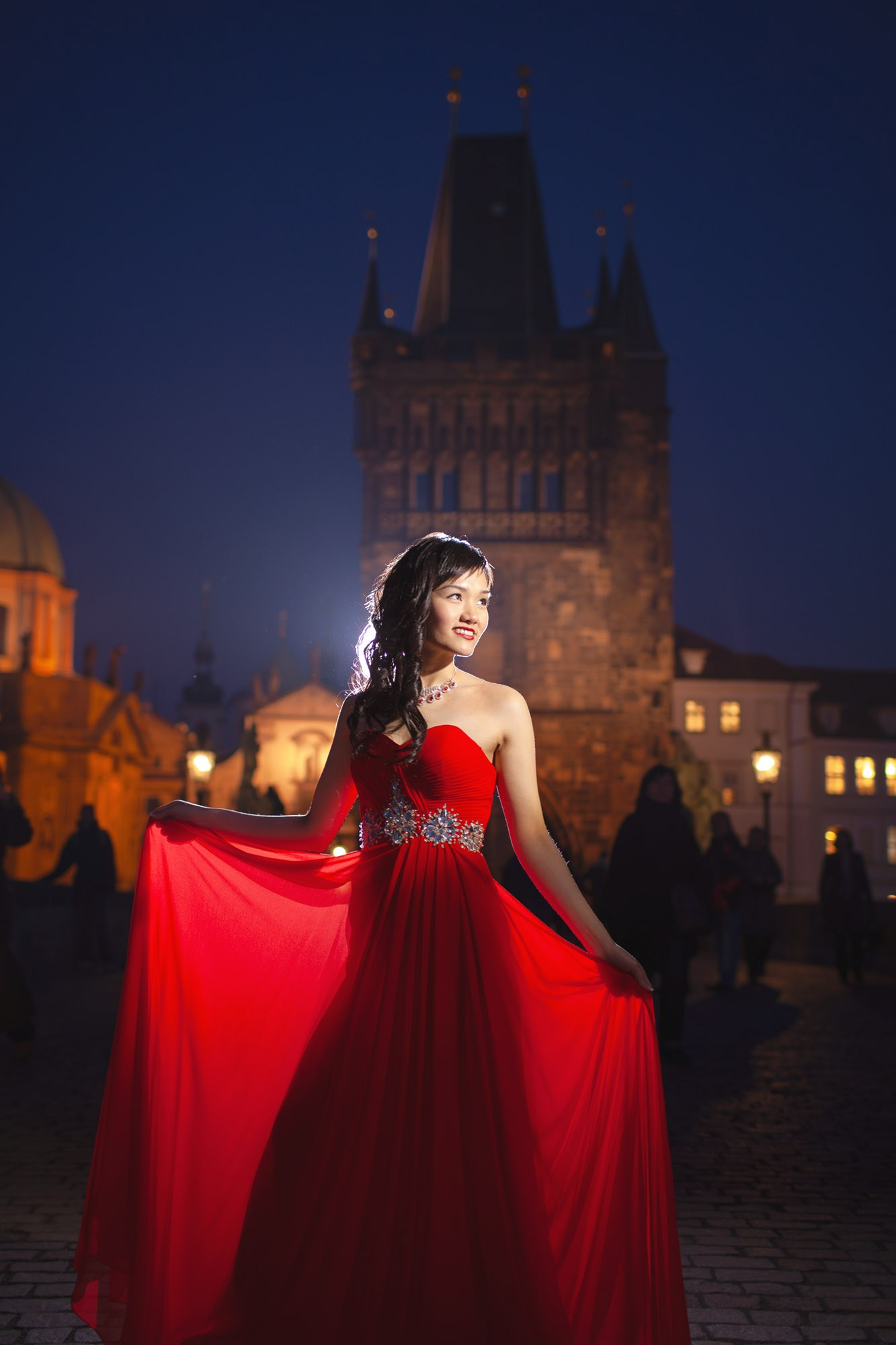 A smiling Hong Kong woman wearing a stylish red evening dress poses for a photo underneath the gas lamps on the historic Charles Bridge at night.