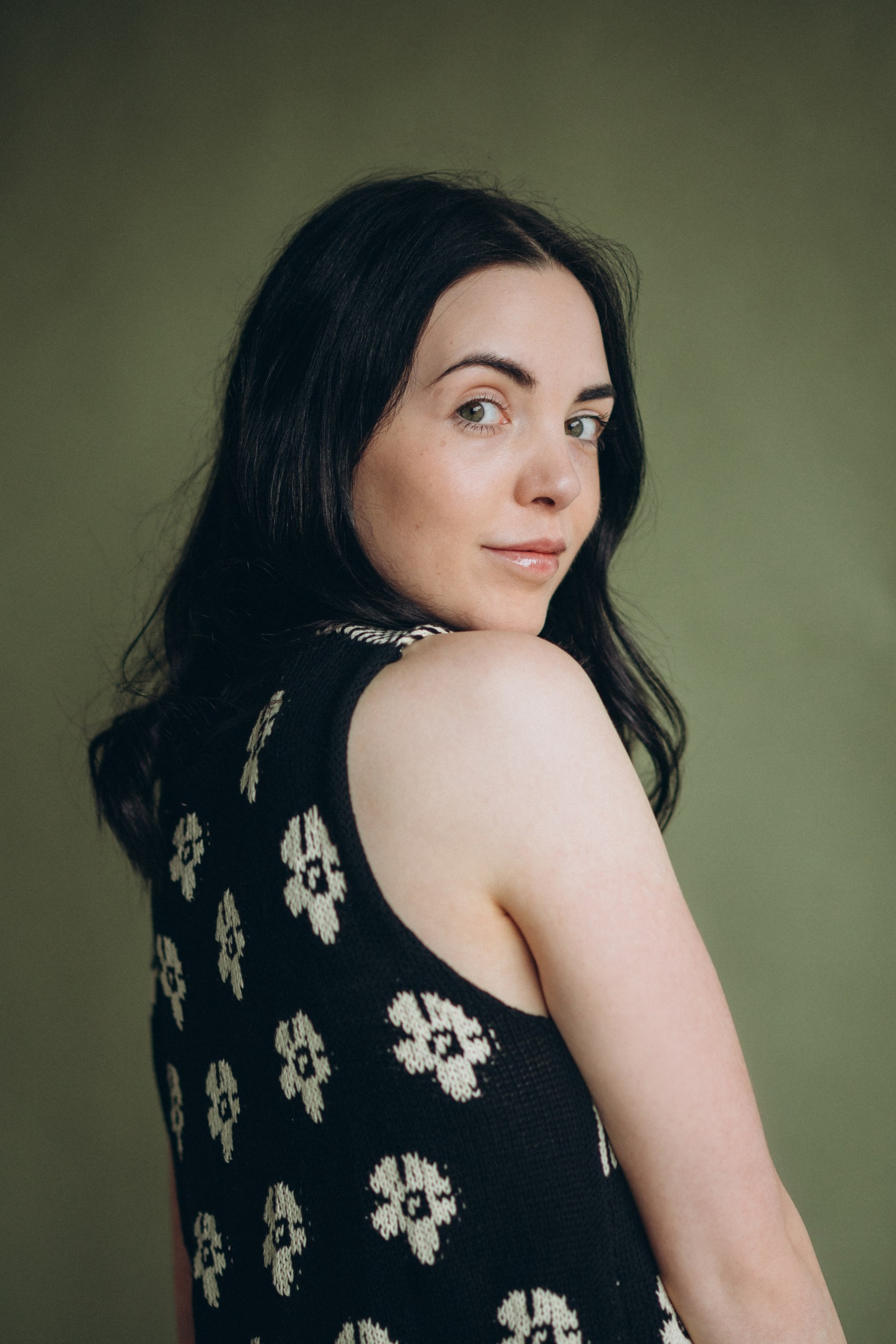 Actor headshot of a woman with long dark hair in a black sleeveless top, looking over her shoulder against a neutral background