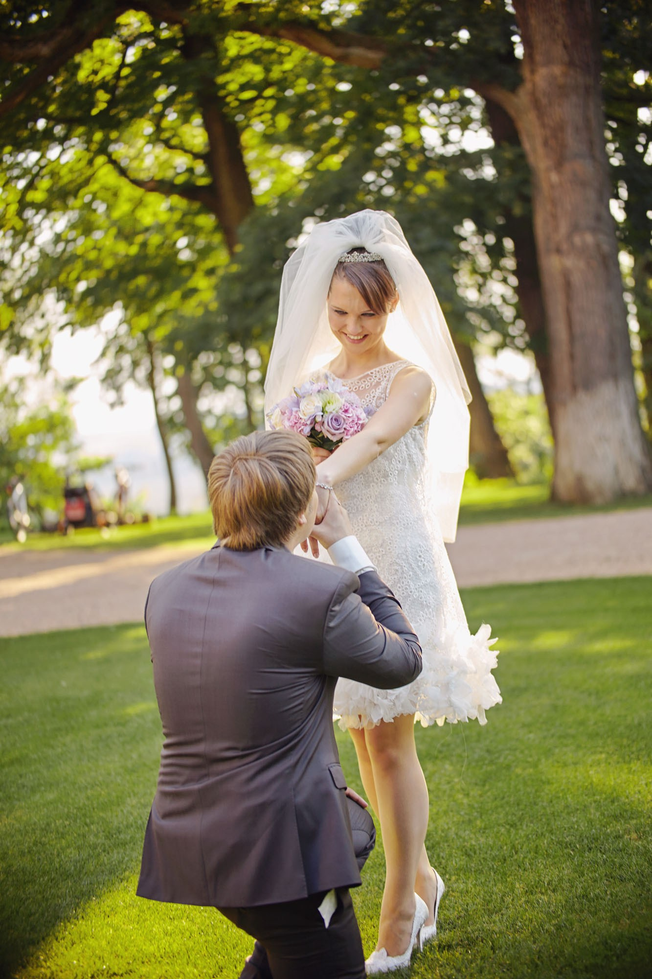 A groom tenderly kisses his radiant bride's hand while kneeling on the picturesque grounds of Chateau Mcely after their romantic outdoor wedding ceremony.