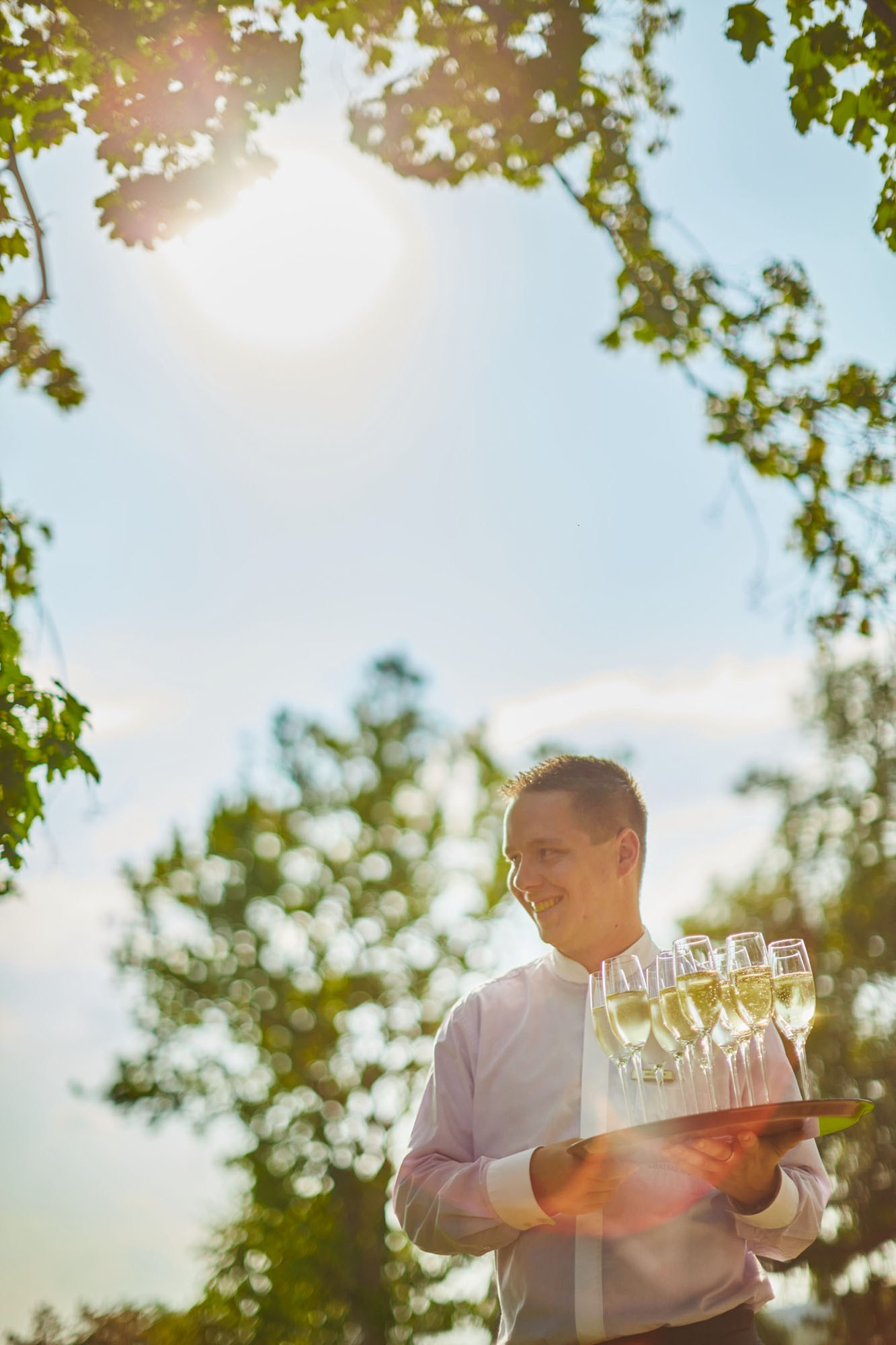 A server holding a try of champagne is backlit by the sun that creates a flare of light on the grounds of the chateau following a wedding.