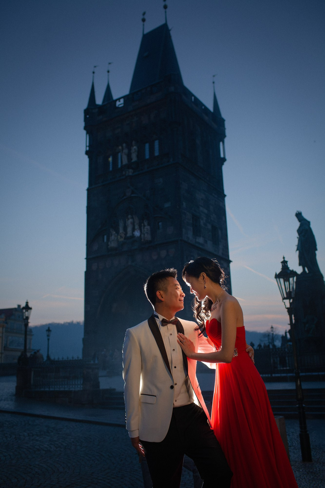 A man wearing a white tuxedo and his bride-to-be wearing a slink red evening dress share a moment near the powder tower near the historic Charles Bridge in Prague.