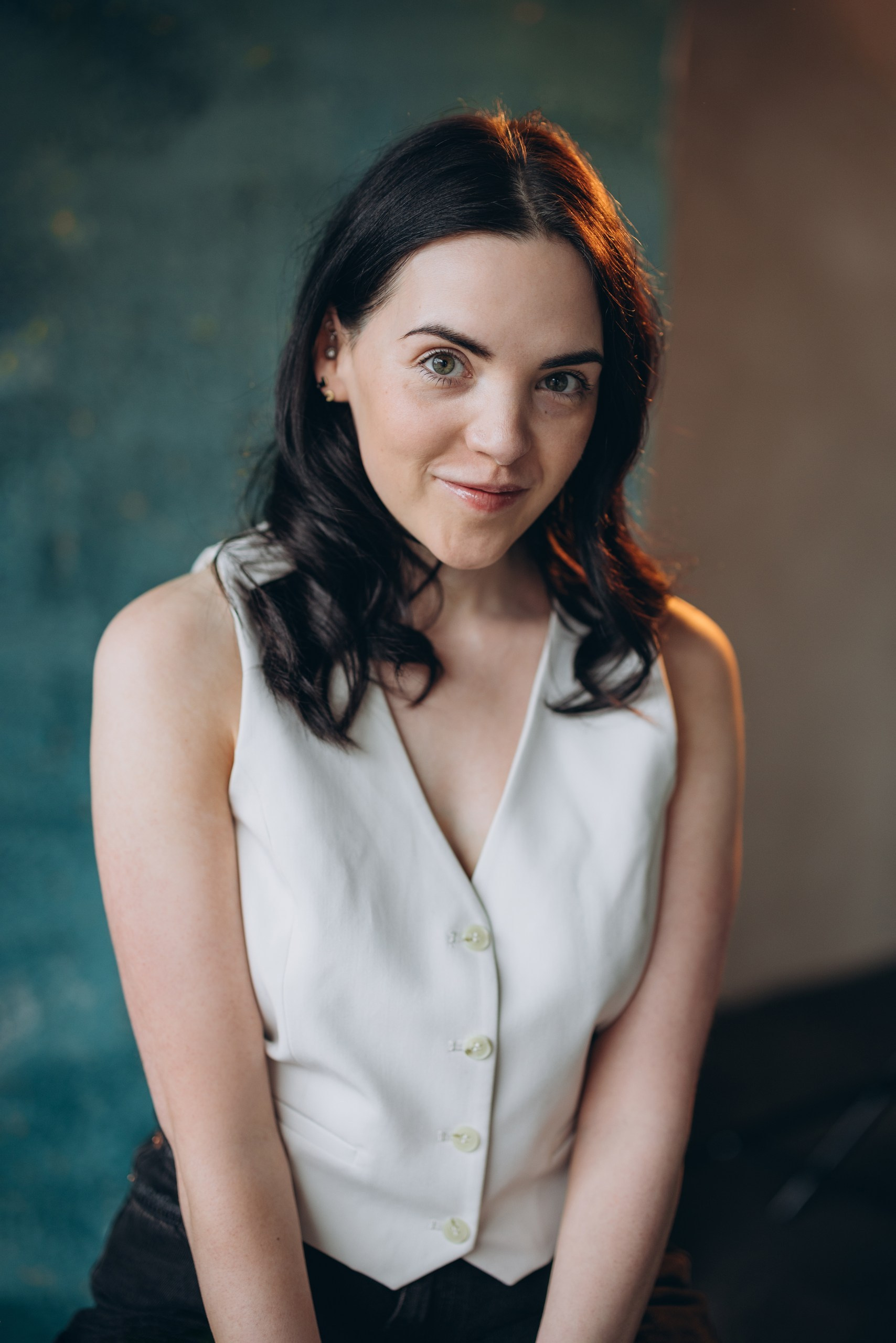 Actor headshot of a smiling woman with dark hair in a white blouse sitting against a blue background