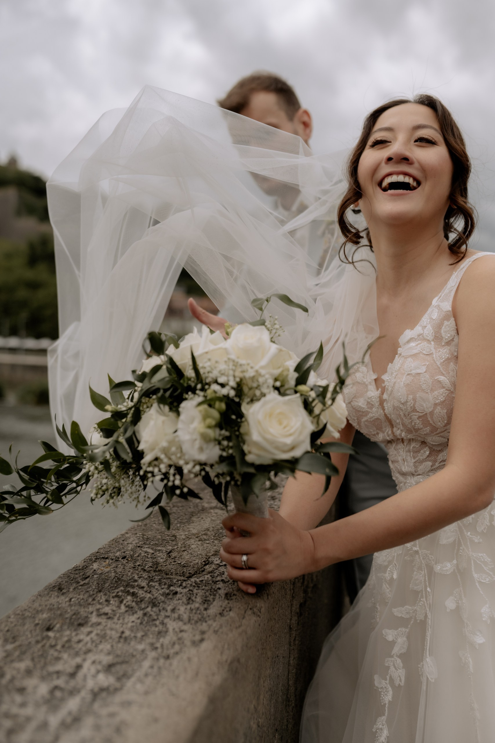 Der Bräutigam hebt seine Braut spielerisch auf der Brücke hoch, beide lachen herzlich – im Hintergrund Menschen, die klatschen und gratulieren