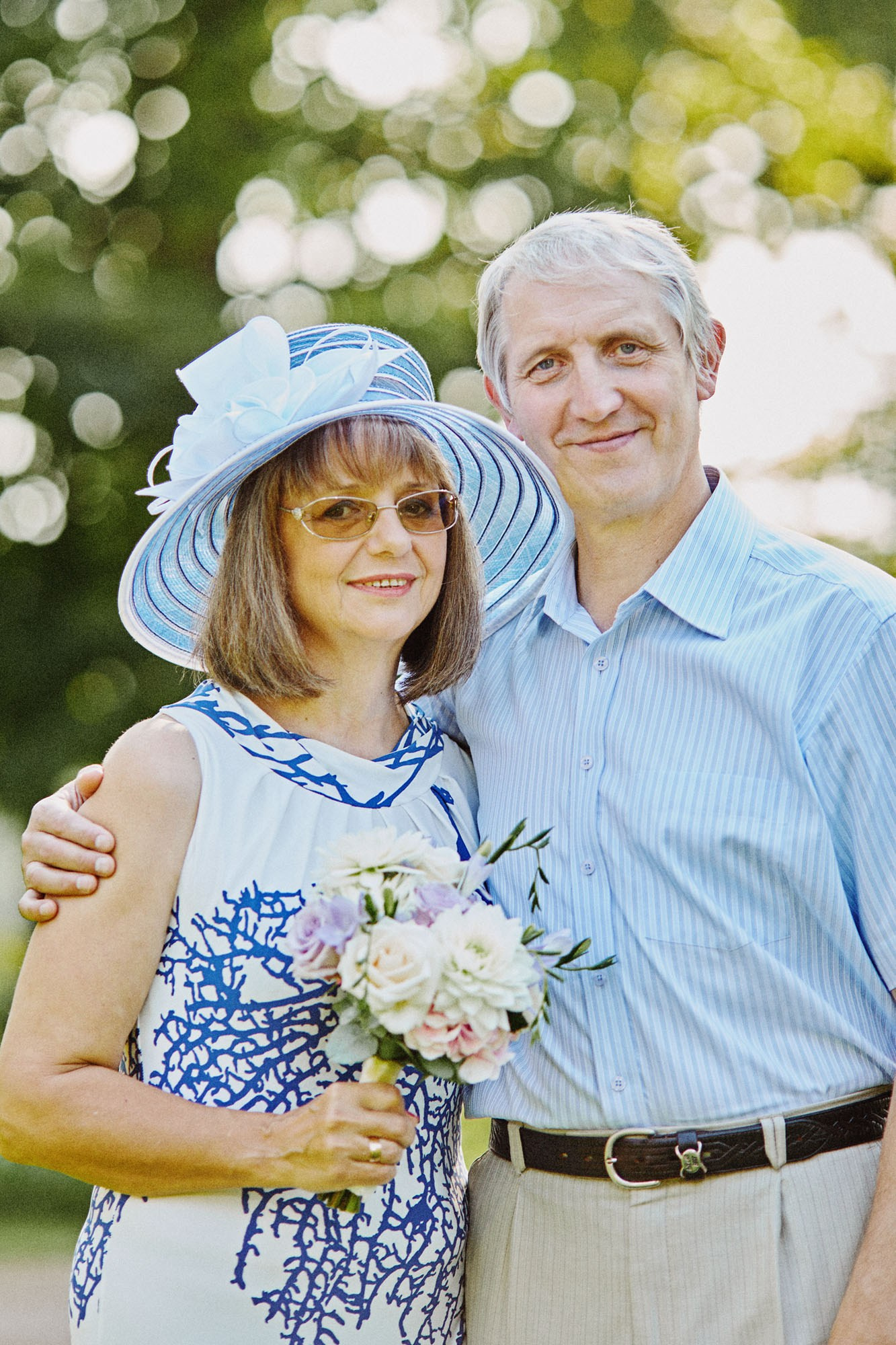Mother of the bride, adorned in a chic blue hat, poses with her husband at their daughter's enchanting summer garden wedding.