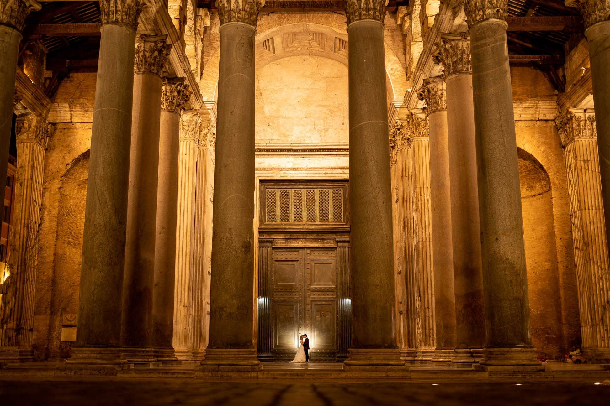 Sposi appena uniti in matrimonio posano di notte davanti al maestoso Pantheon illuminato a Roma, immortalati da un fotografo matrimonio professionista. L'immagine cattura un momento romantico e senza tempo, con la coppia al centro e la storica architettura che fornisce uno sfondo suggestivo, esemplificando l'eccellenza nella fotografia di matrimonio.