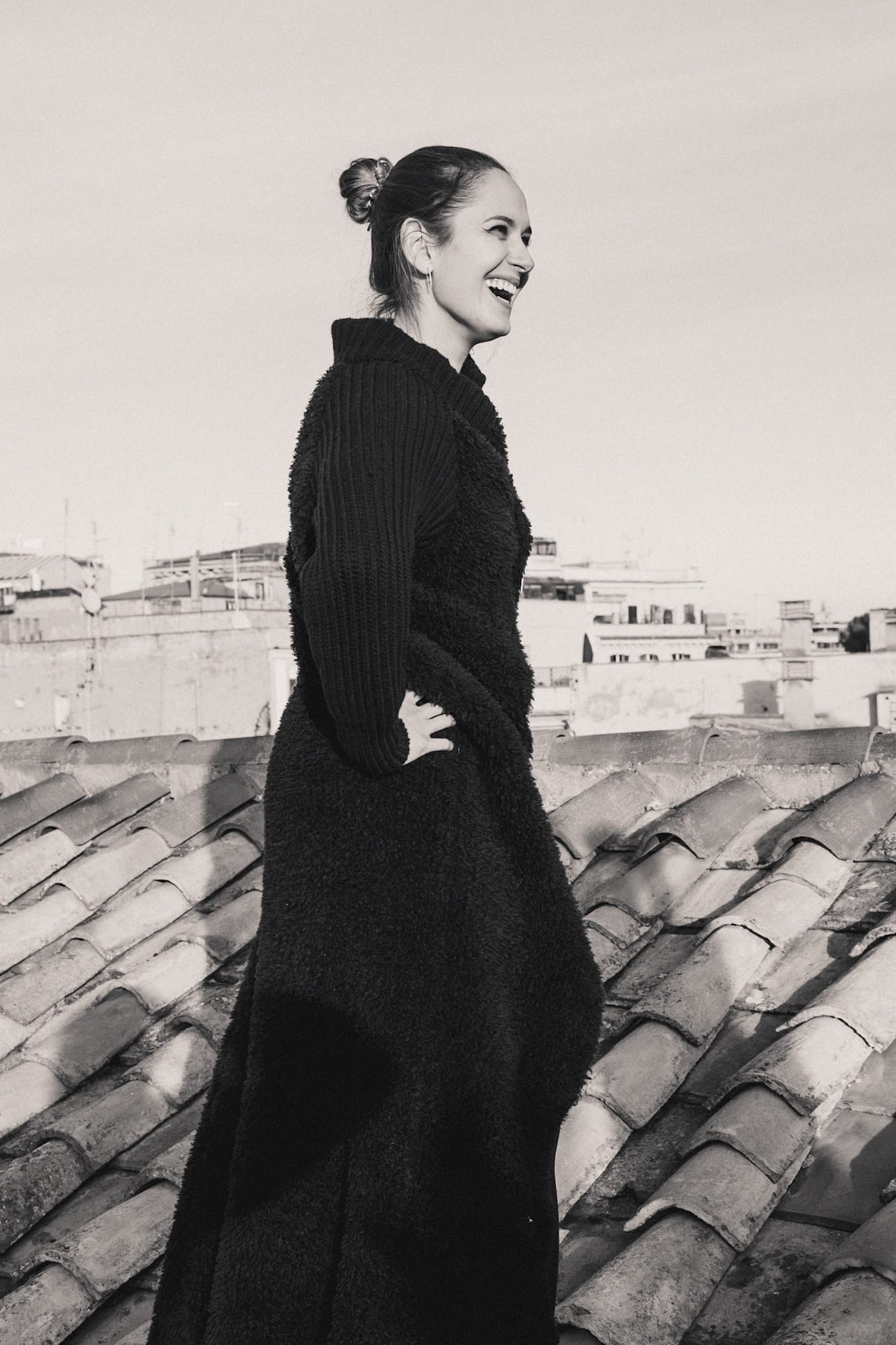 A lifestyle photography of a beautiful young woman in a long black coat in Rome laughing on the rooftop.