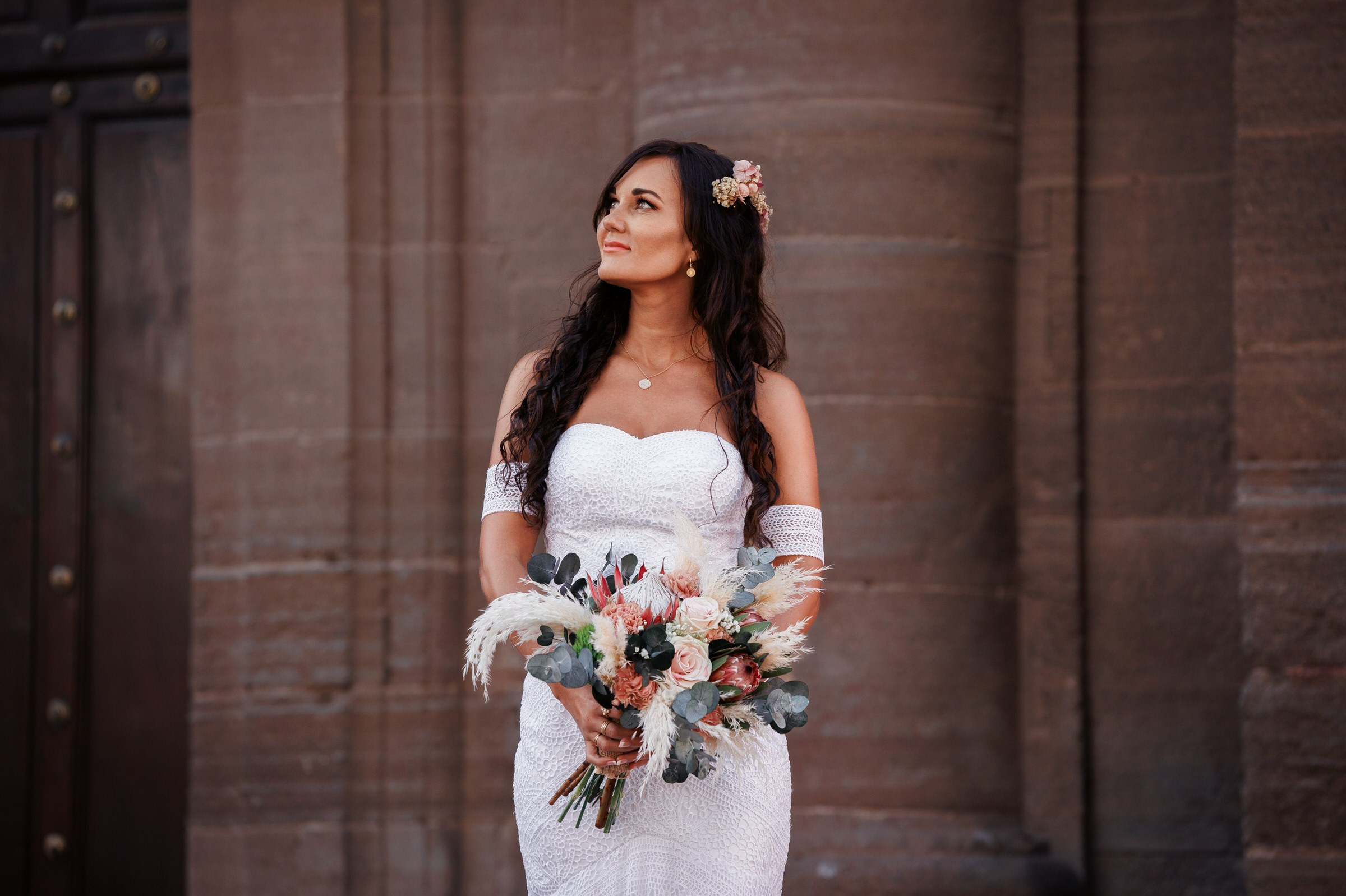 Une femme vêtue d'une robe de mariée blanche à épaules dénudées tient un bouquet de fleurs et regarde vers le haut, debout devant un mur de pierre.
