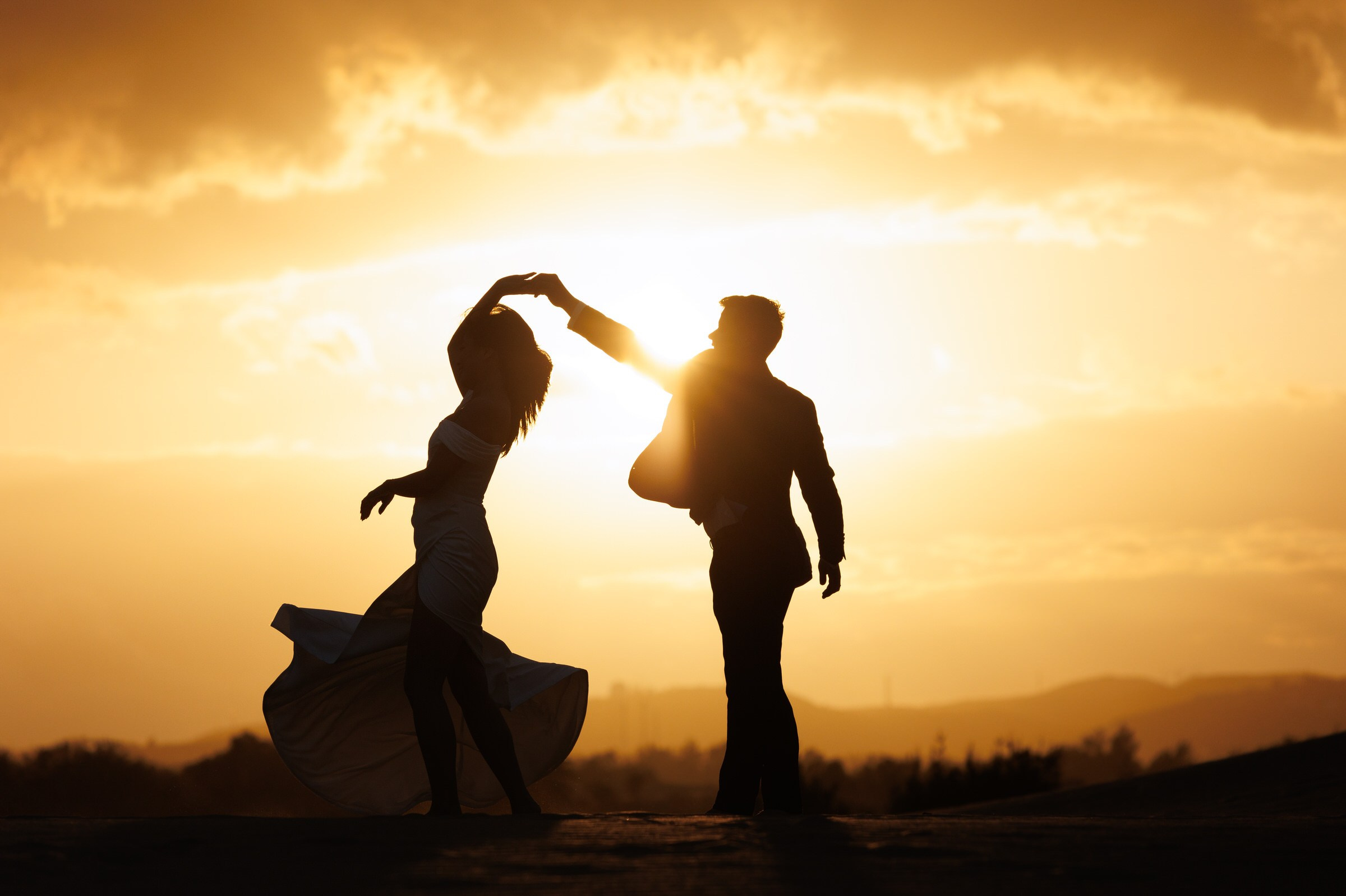 Silhouettes d'un homme et d'une femme dansant à l'extérieur au coucher du soleil avec le soleil bas dans le ciel derrière eux.