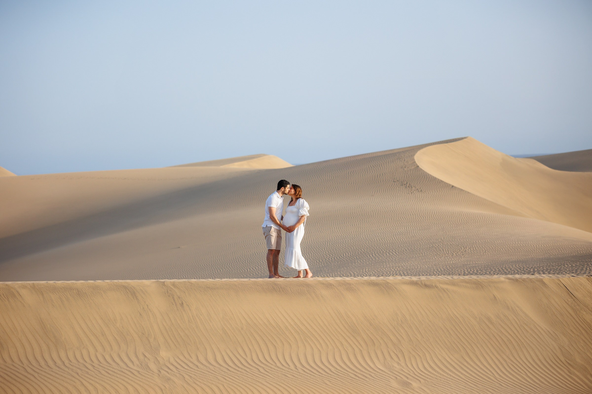 Un couple se tient face à face sur une dune de sable dans un paysage désertique sous un ciel clair.
